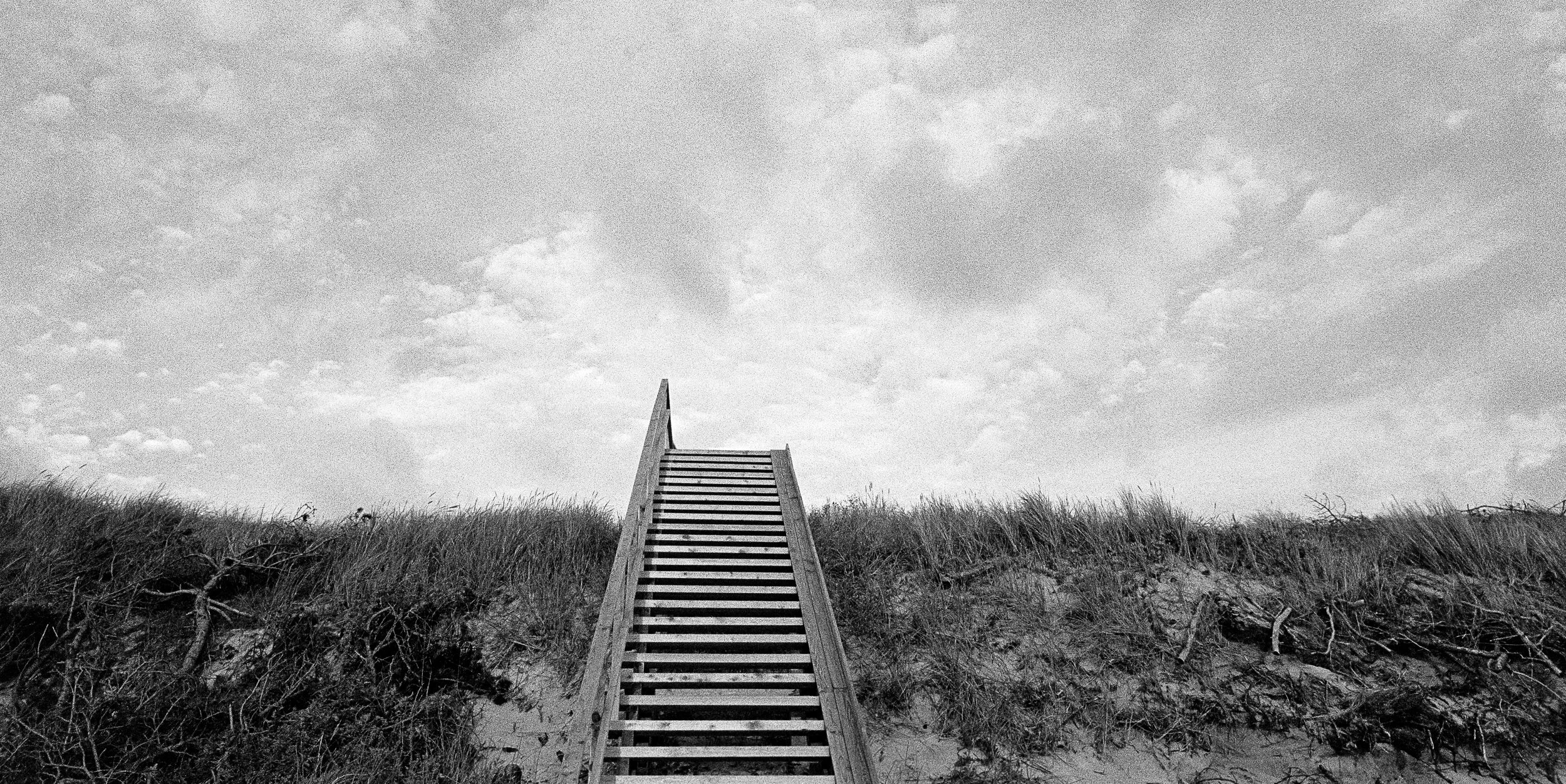 Minimalist black and white analog photograph of a wooden staircase rising through sand dunes toward an open sky by Donatas Berlinskas
