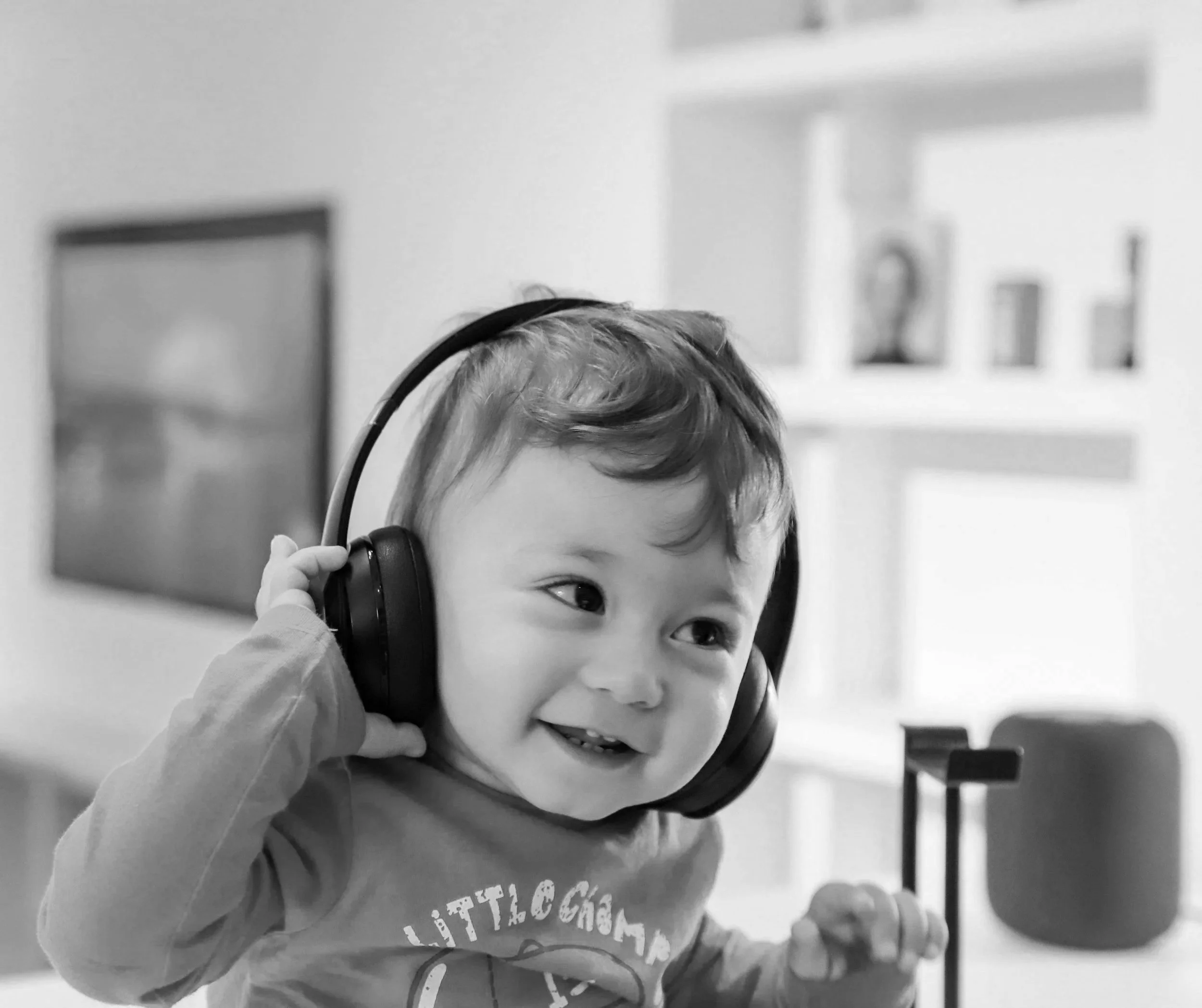 A young child with curly hair wearing headphones and smiling while looking to the side indoors.