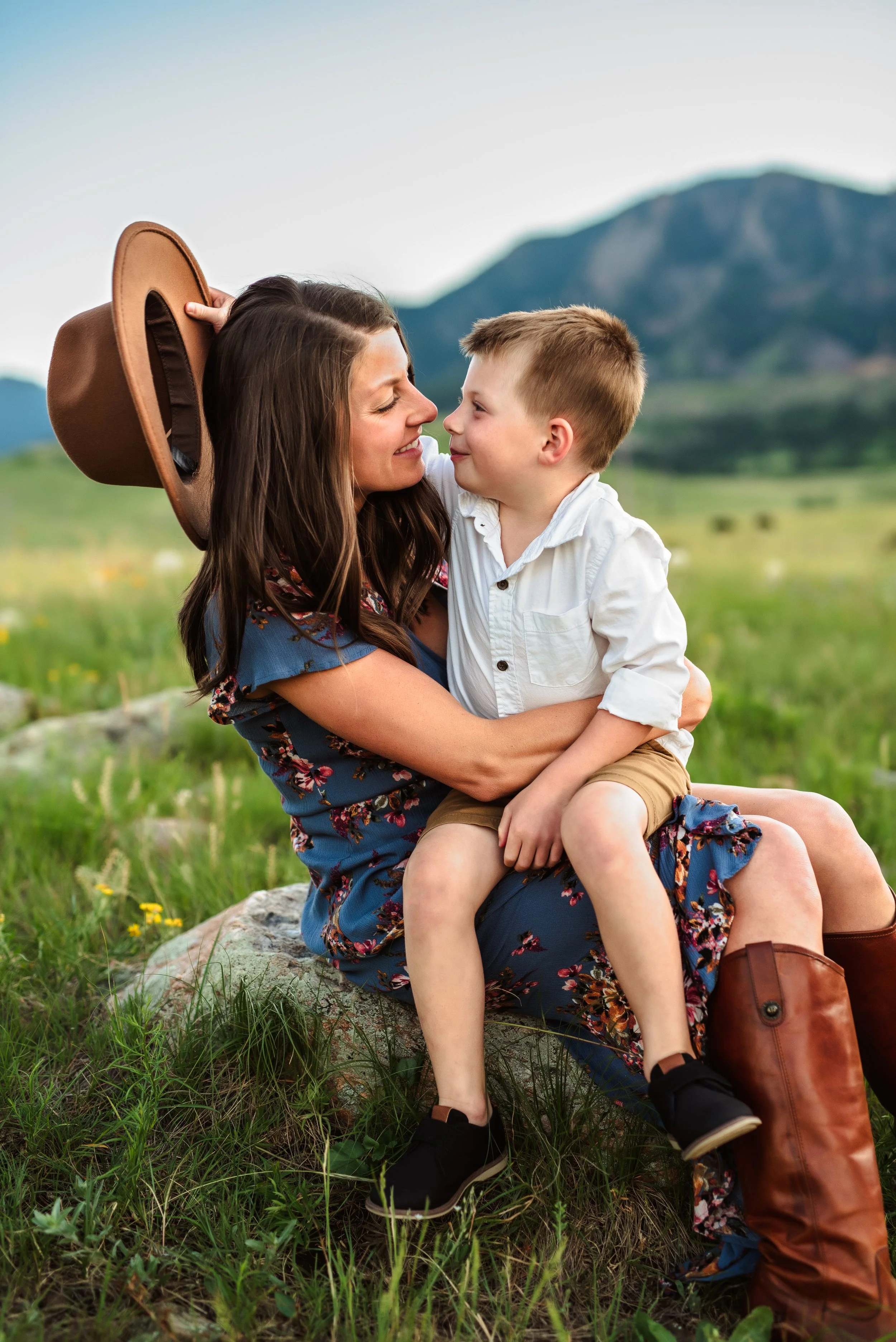 A woman and a young boy sitting on a rock outdoors in a grassy field with mountains in the background. The woman is smiling at the boy, who is holding her face and smiling back. The woman has long brown hair and is wearing a floral dress, and the boy has short blonde hair and is wearing a white shirt and shorts. The woman has a large hat resting on her head, and there is a brown backpack on the ground next to her.