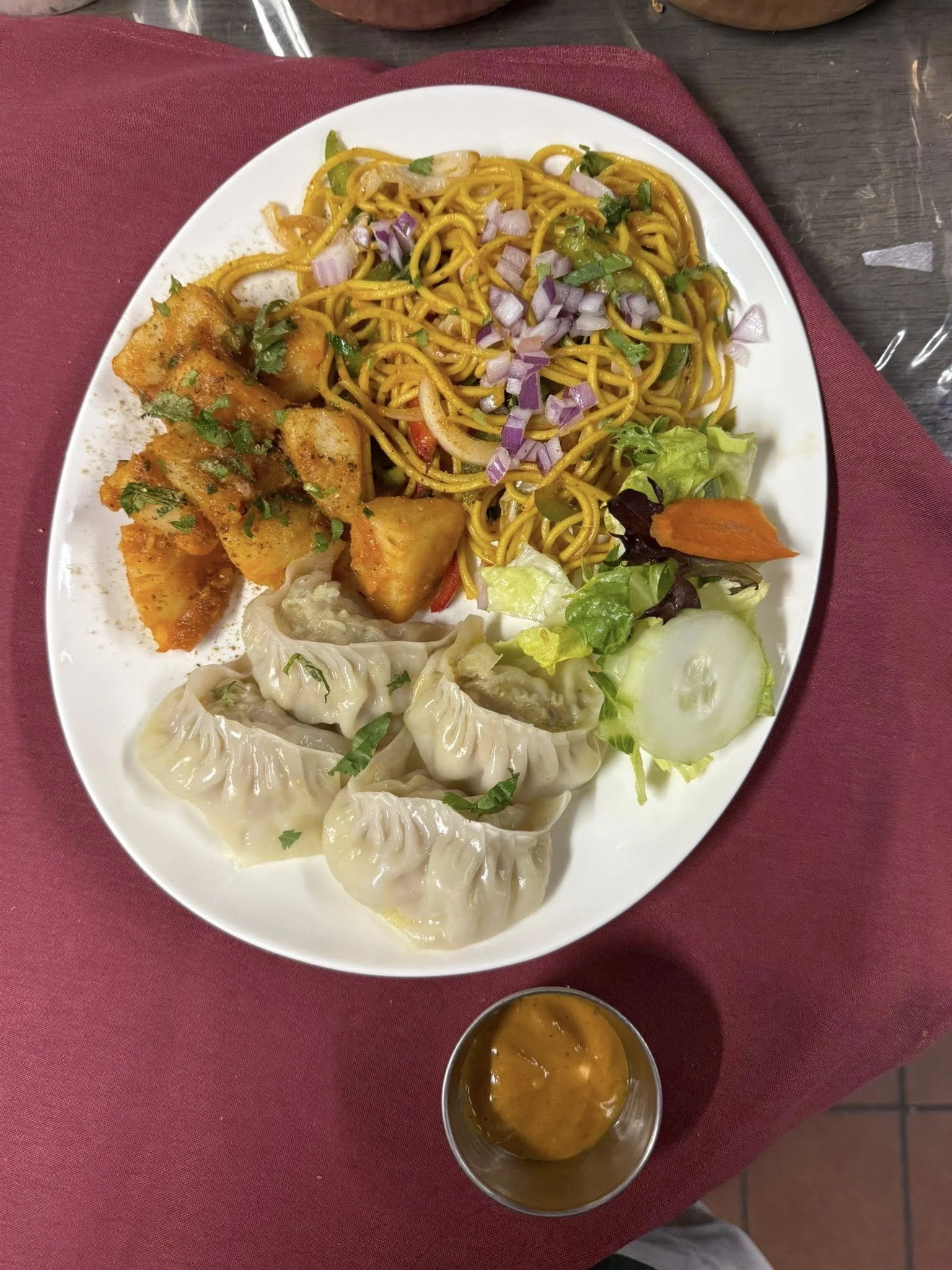 A white plate with Indian food, including fried dumplings, noodles with vegetables, and vegetable curry, accompanied by a small metal container of sauce, set on a red tablecloth.