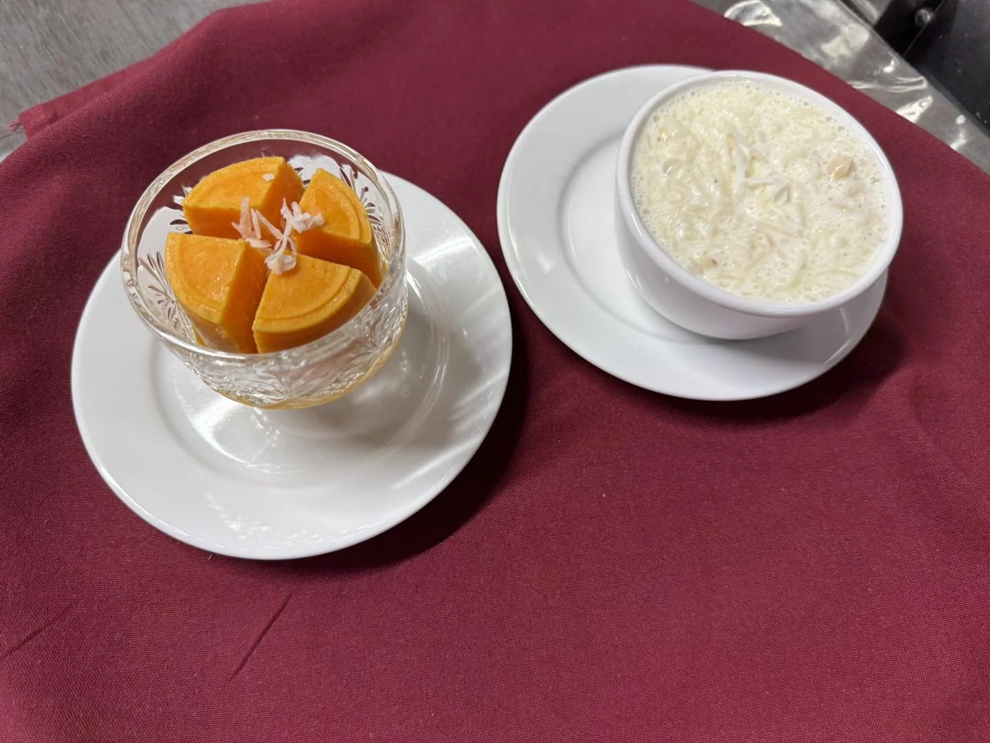 Glass bowl with orange slices topped with shredded coconut, and a cup of Indian dessert, kheer, on a red tablecloth.