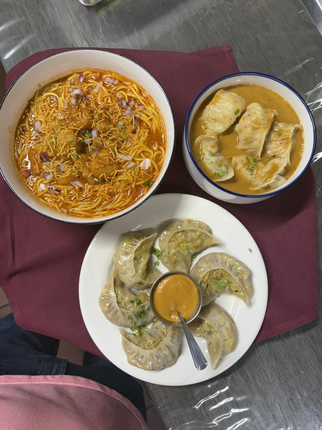 Top-down view of Indian food including a bowl of spicy noodle soup, a bowl of dumplings in yellow curry, and a plate of steamed dumplings with dipping sauce.