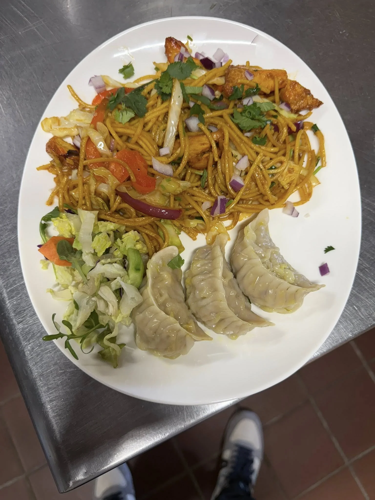 A white oval plate with Asian food including stir-fried noodles with vegetables and tofu, four steamed dumplings, and a side salad with lettuce, cucumber, tomato, and shredded carrots.