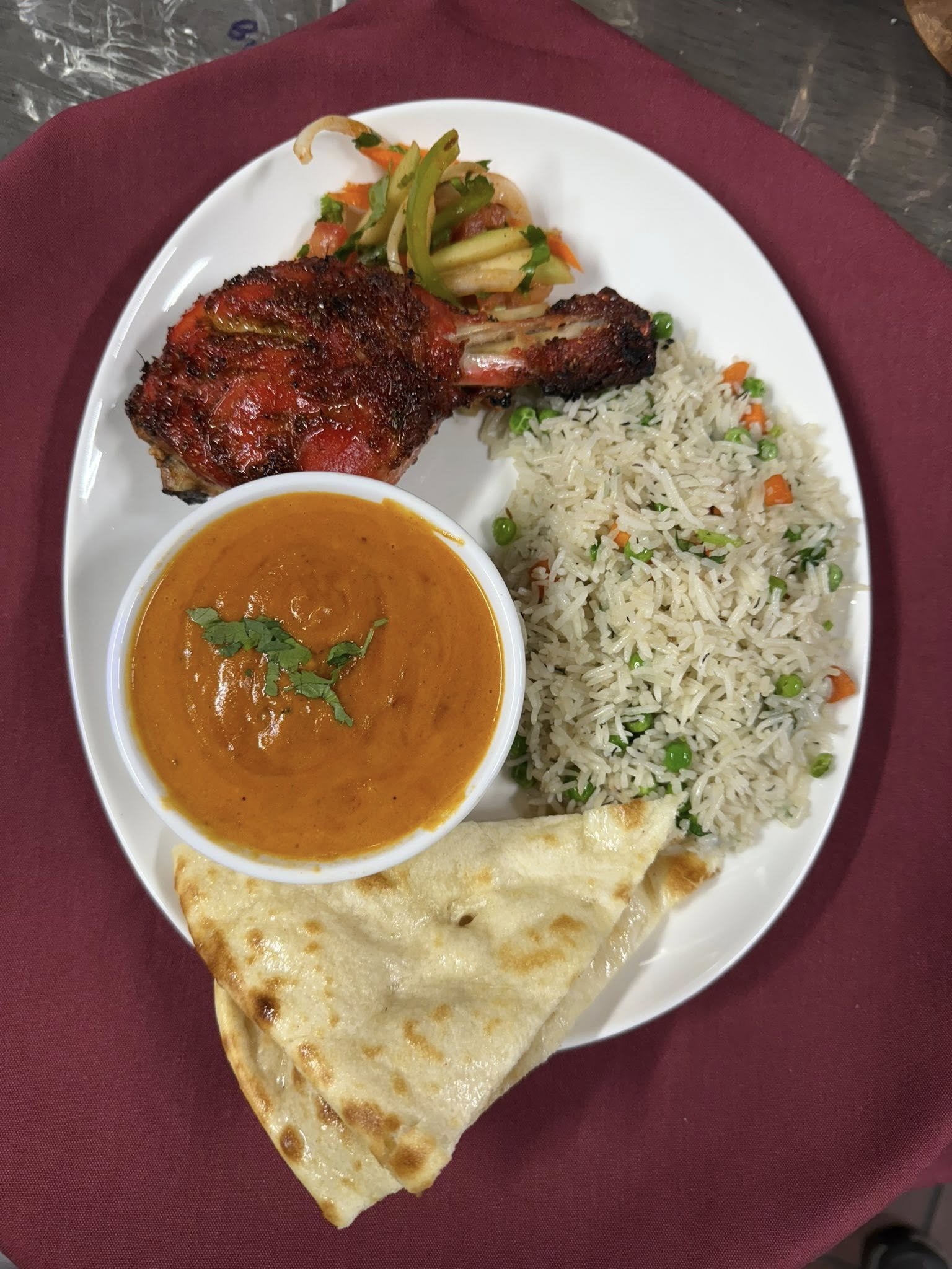 An Indian meal on a white plate includes grilled chicken, vegetable salad, rice with peas and carrots, a bowl of curry, and a piece of naan bread.