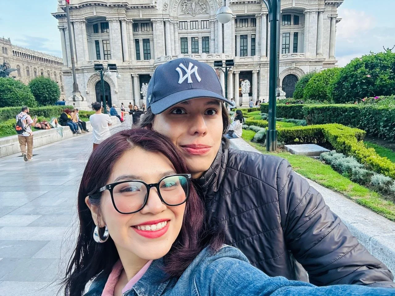 Dos personas sonriendo y tomándose una selfie frente a un edificio histórico con jardines verdes.