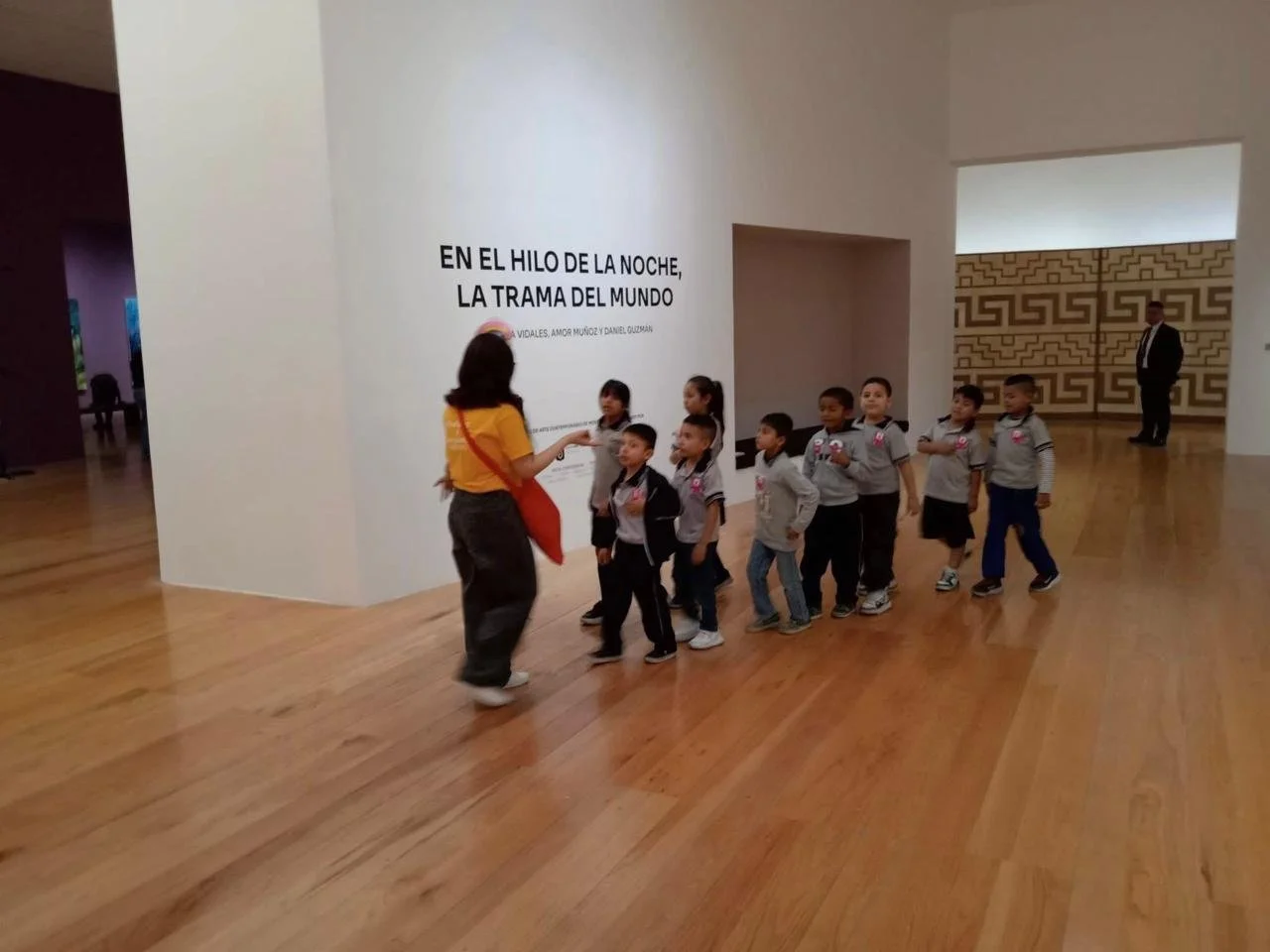 Grupo de niños en una visita escolar guiada por una mujer en un museo, con un adulto vigilando en el fondo.