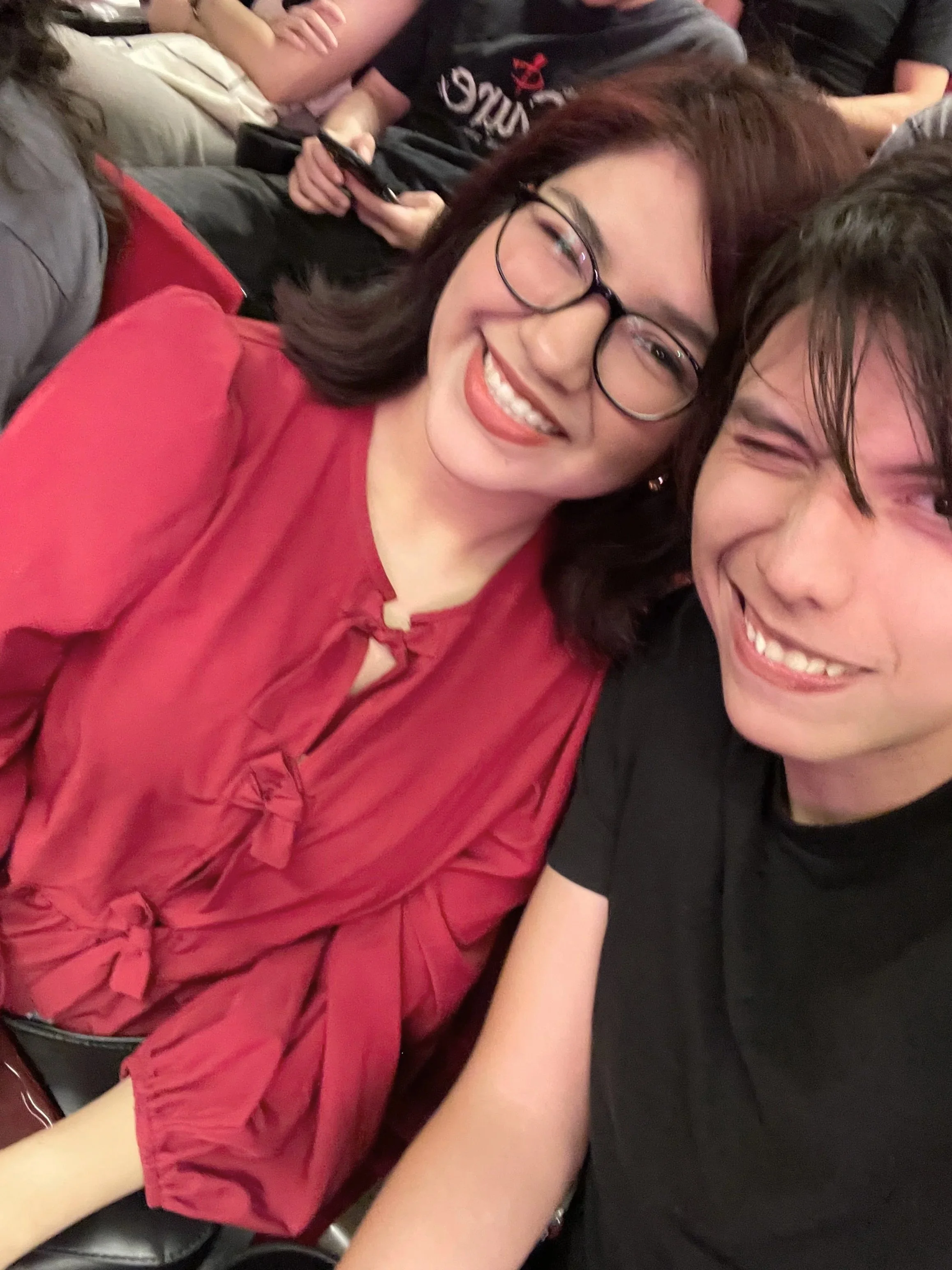 Dos personas sonrientes, una mujer con gafas y cabello rizado, vestida con blusa roja, y un joven con cabello oscuro y camiseta negra, tomándose una selfie en un ambiente social. En el fondo, otras personas parecen estar en una reunión.