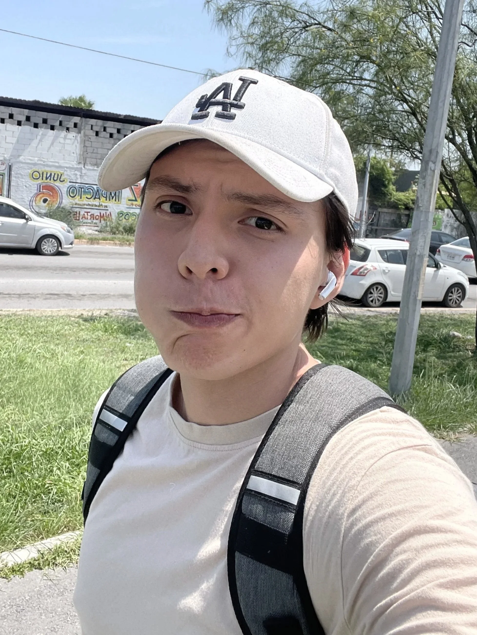 Un joven con gorra blanca con letras negras, camiseta beige y mochila gris tomando una selfie en la calle, con autos y árboles en el fondo.
