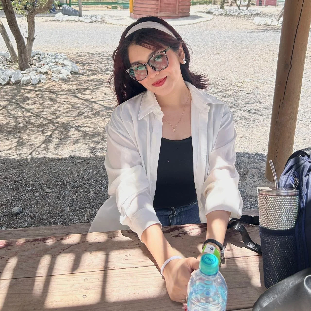 Mujer con gafas y blusa blanca, sonriendo, sentada en una mesa de madera, en un lugar al aire libre con sombra y árboles.