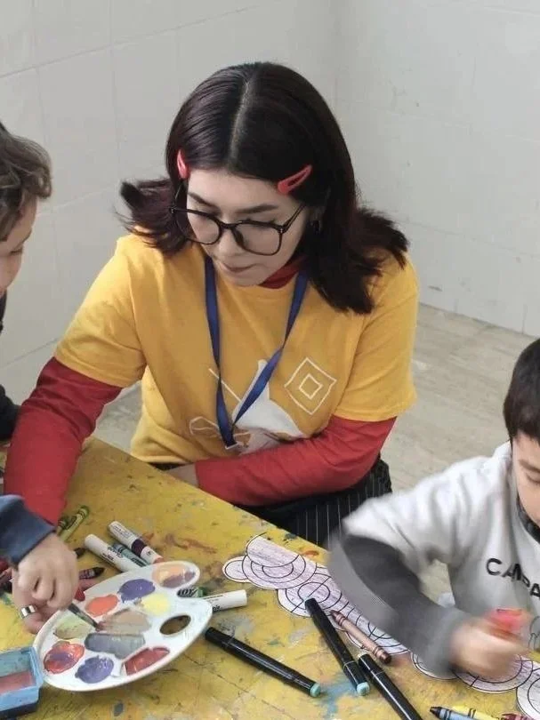 Mujer con gafas y cabello oscuro, usando camiseta amarilla, enseña a niños a pintar en una mesa con marcadores y paleta de colores.