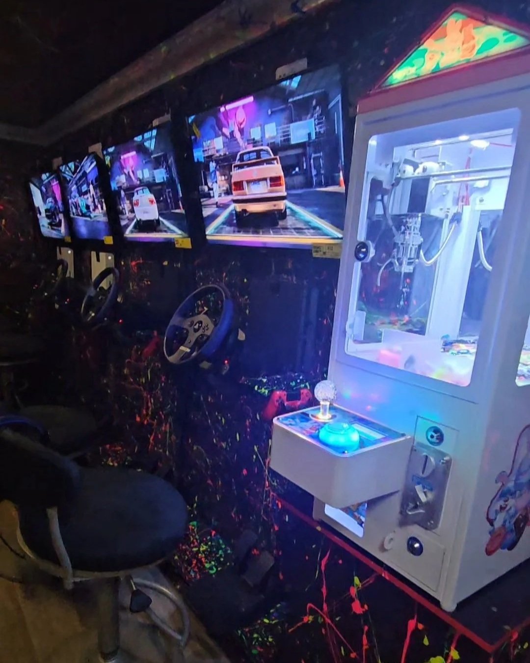 Four arcade racing game screens showing cars on a city street, with multiple steering wheels in front of each screen, and a claw machine to the right with a glowing blue orb inside.