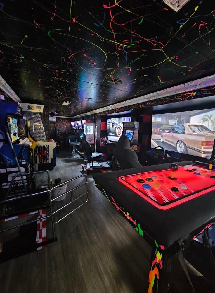 An arcade room with multiple gaming machines, a colorful LED-lit arcade controller on a table, and cars visible through large windows.