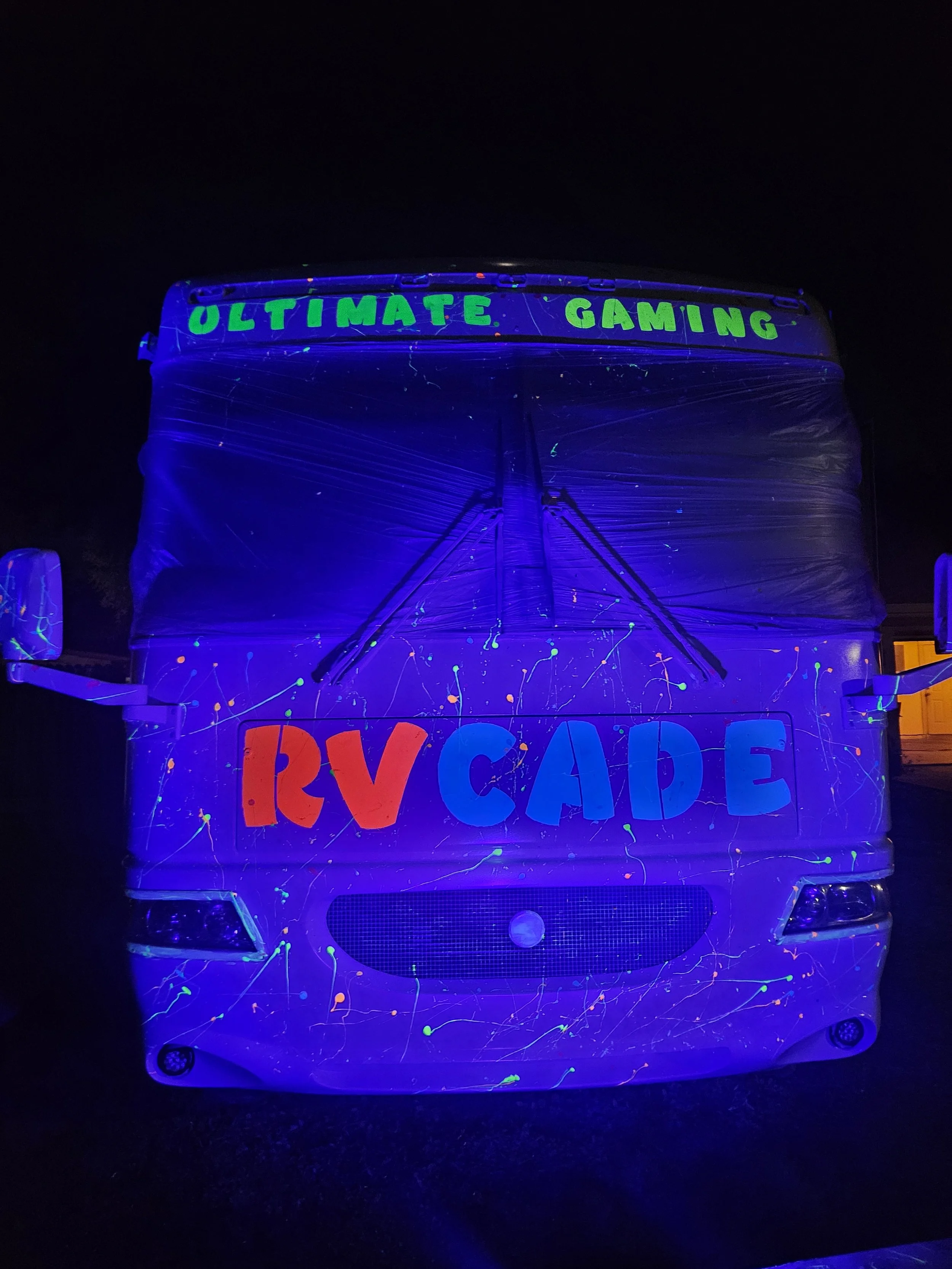 Front of a glowing bus with neon paint in blacklight, displaying "ULTIMATE GAMING" at the top and "RV CAFE" in large colorful letters on the front.