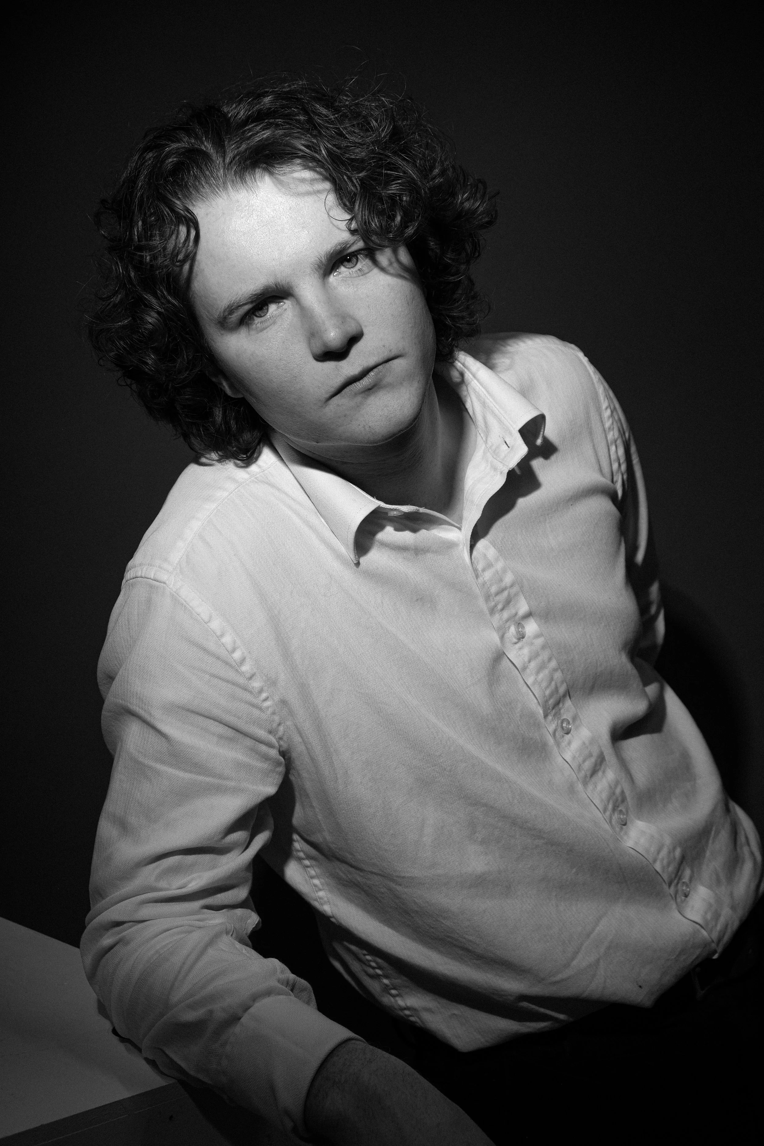 Black and white photo of a young man with curly hair wearing a button-up shirt, looking directly at the camera with a serious expression.
