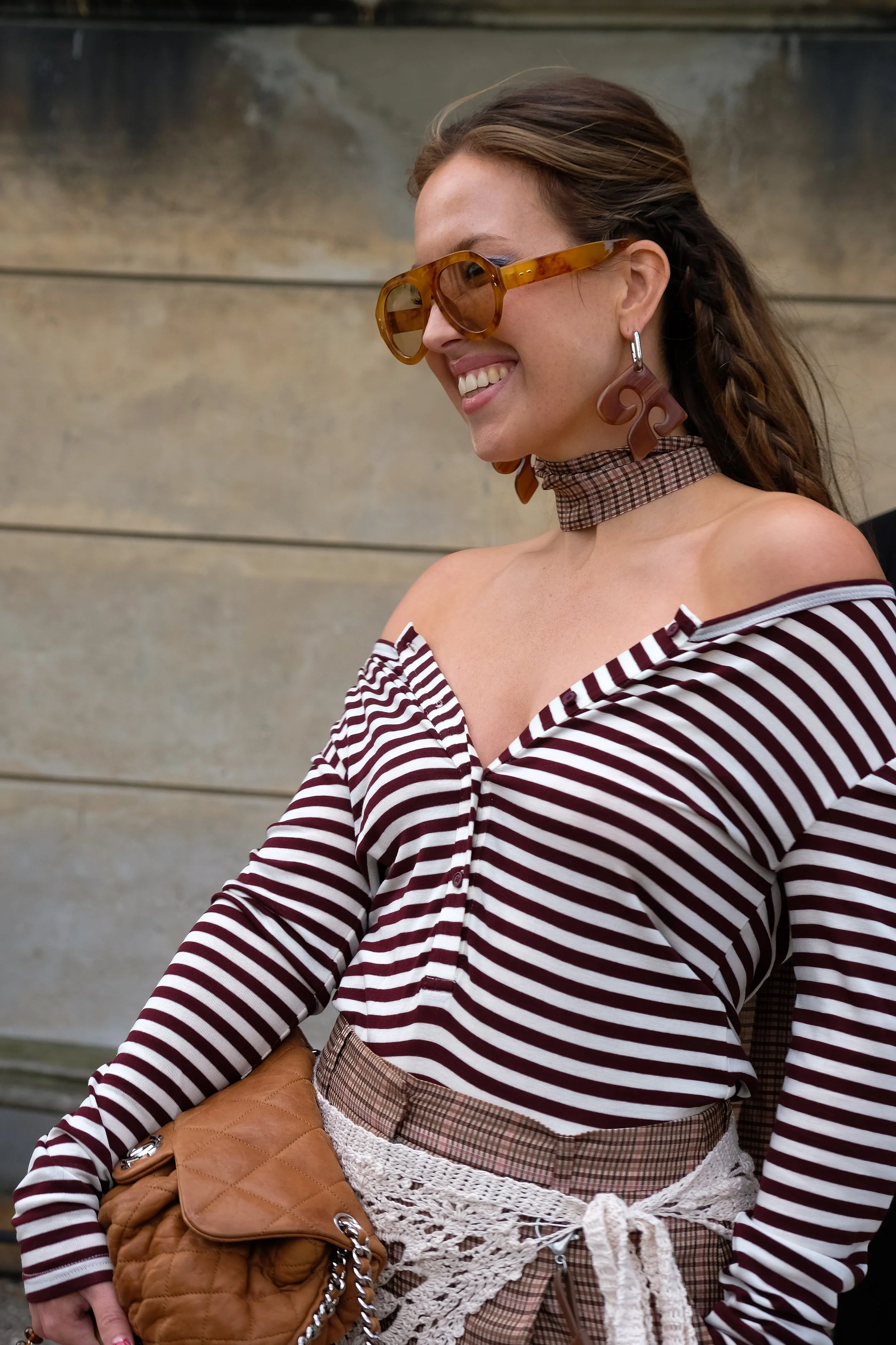A woman wearing large tortoiseshell sunglasses, striped off-the-shoulder top, plaid skirt with a crocheted belt, and chunky earrings, smiling and looking to the side.
