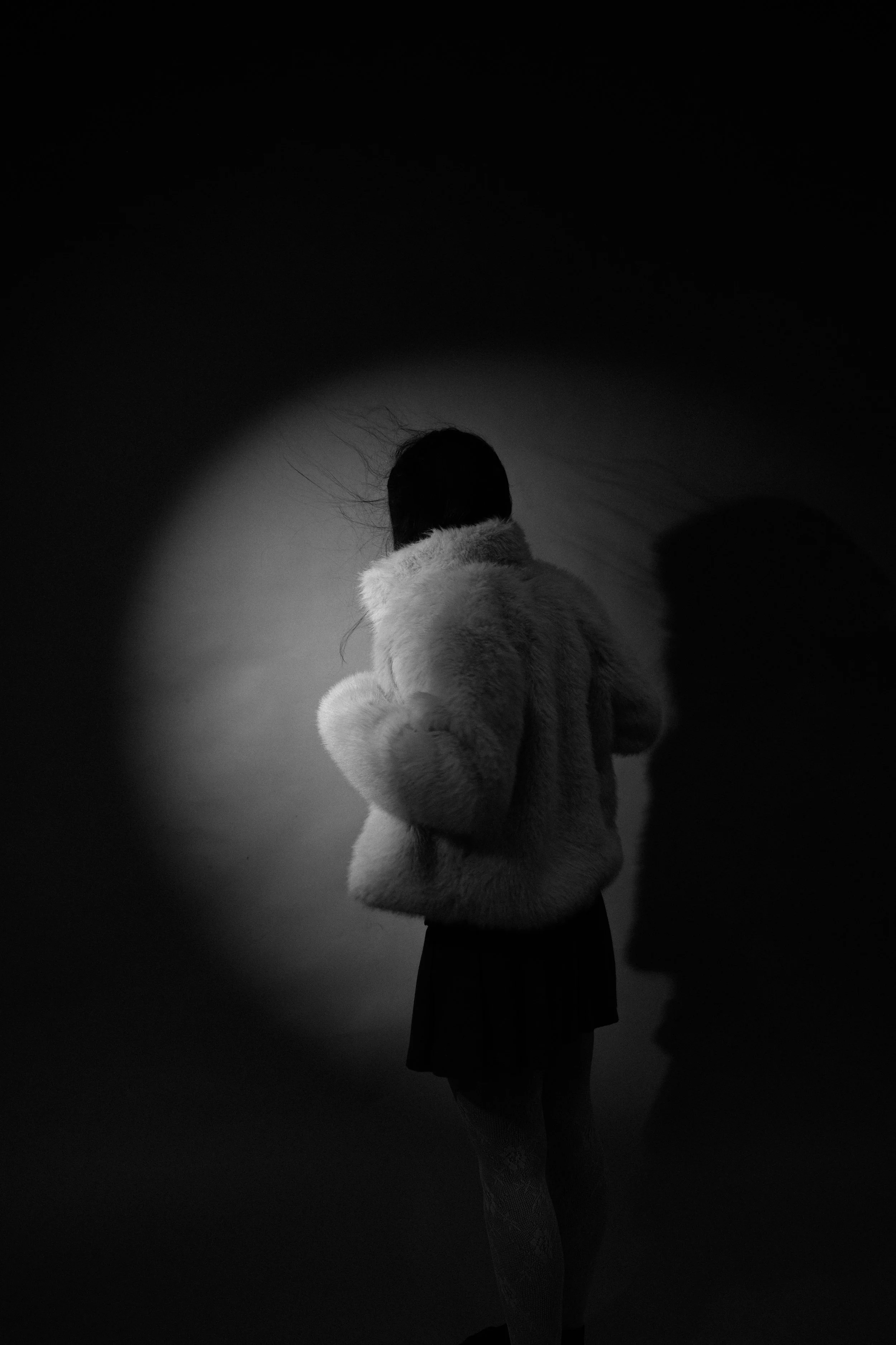 A person wearing a fluffy coat and a skirt, standing with their back facing the camera in a dark room, illuminated by a spotlight, with their hair blowing, and their shadow visible on the wall.