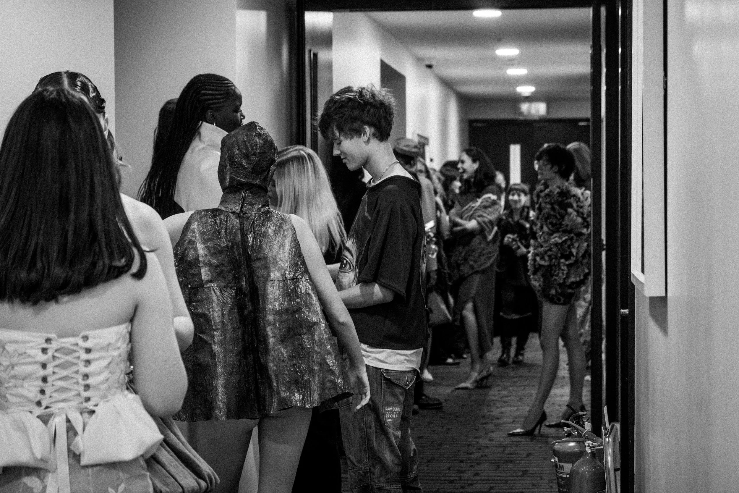 People lined up in a hallway at an indoor event, with some dressed in costumes, including a person in a shiny hooded cape and others in decorative or fashionable outfits.