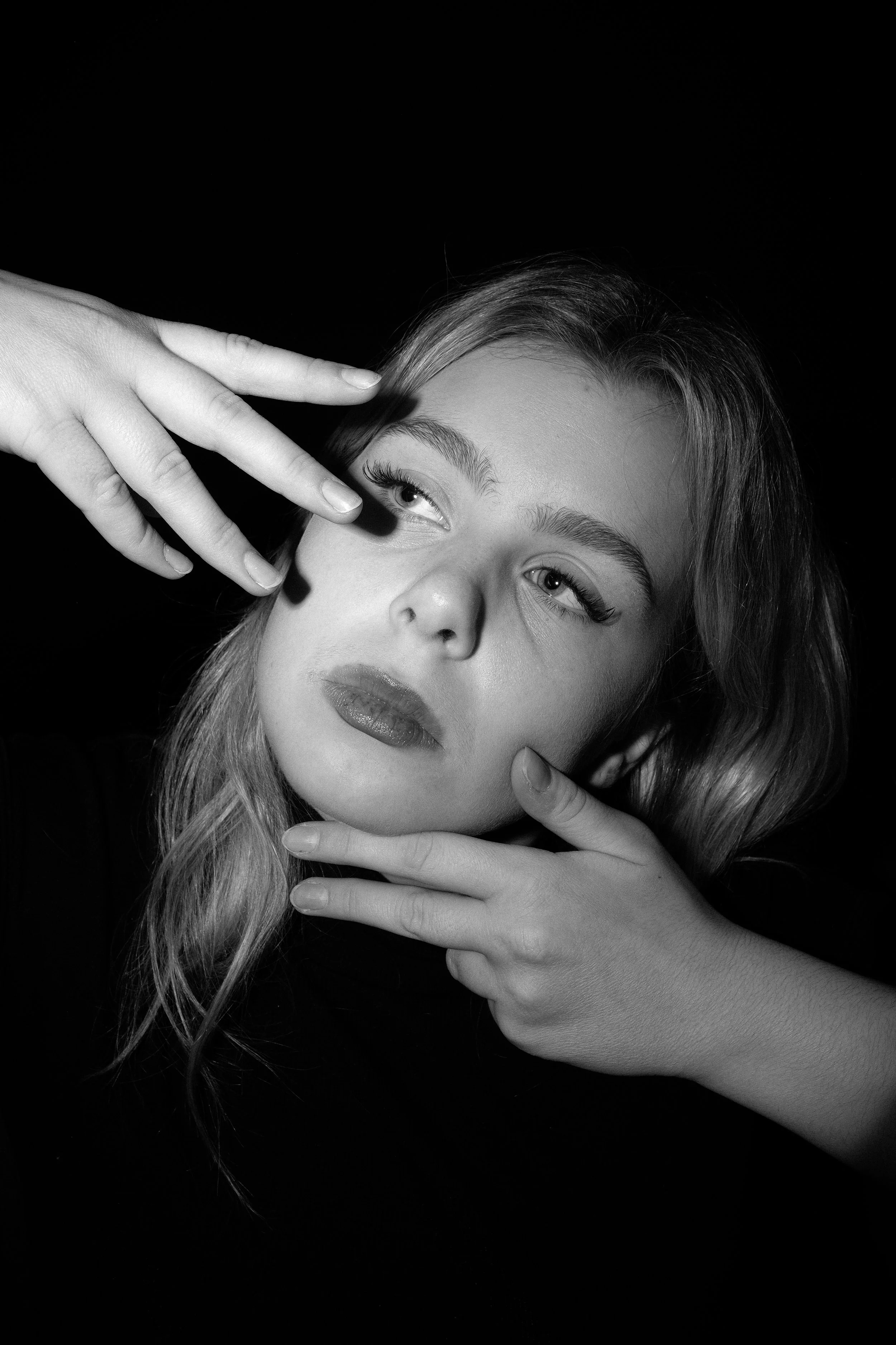 A woman with long hair and makeup posing against a dark background, with her hands near her face.
