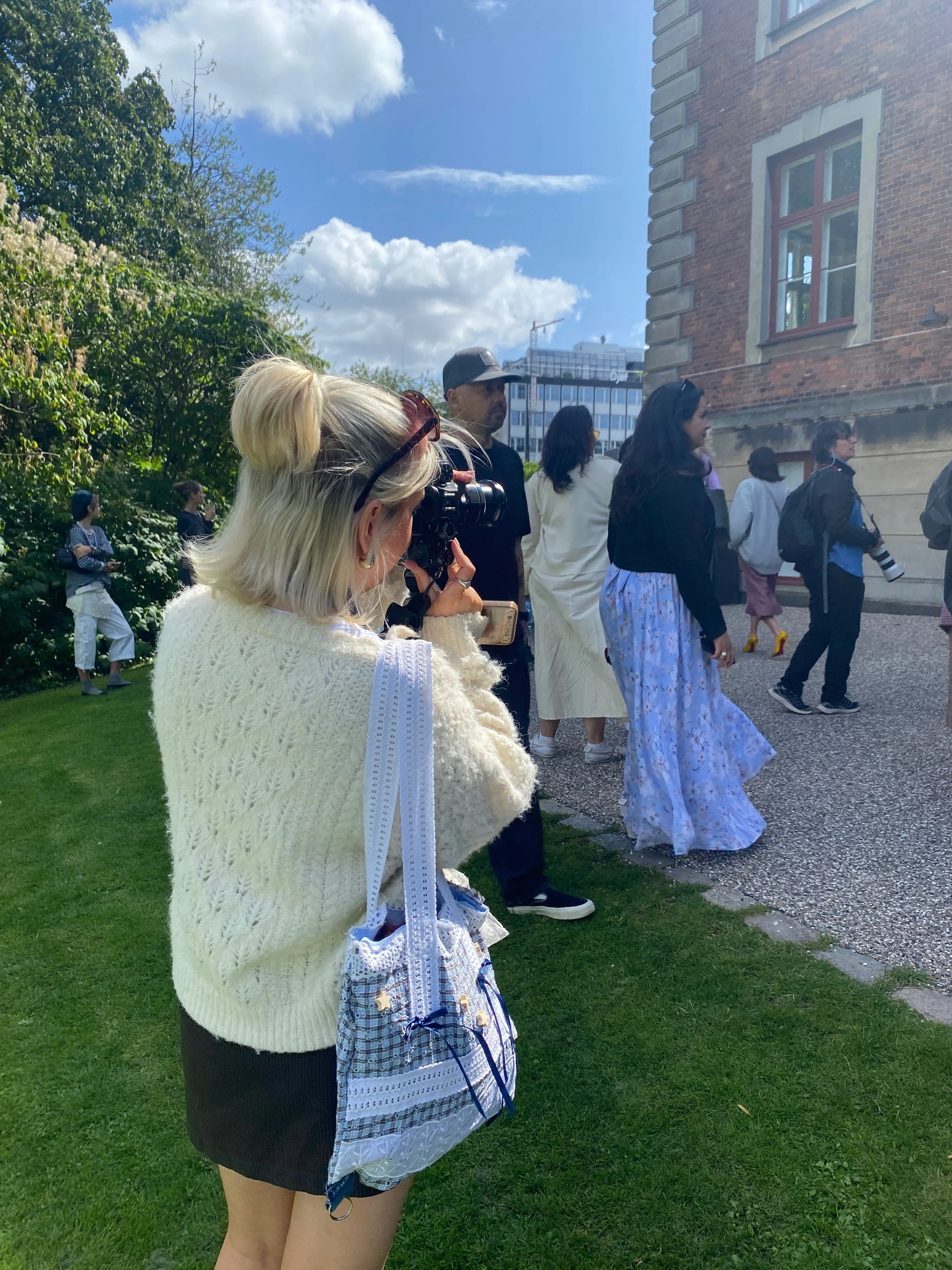 A group of people standing outside near a brick building on a sunny day, with some individuals holding cameras, and a woman in the foreground taking a photo or recording.