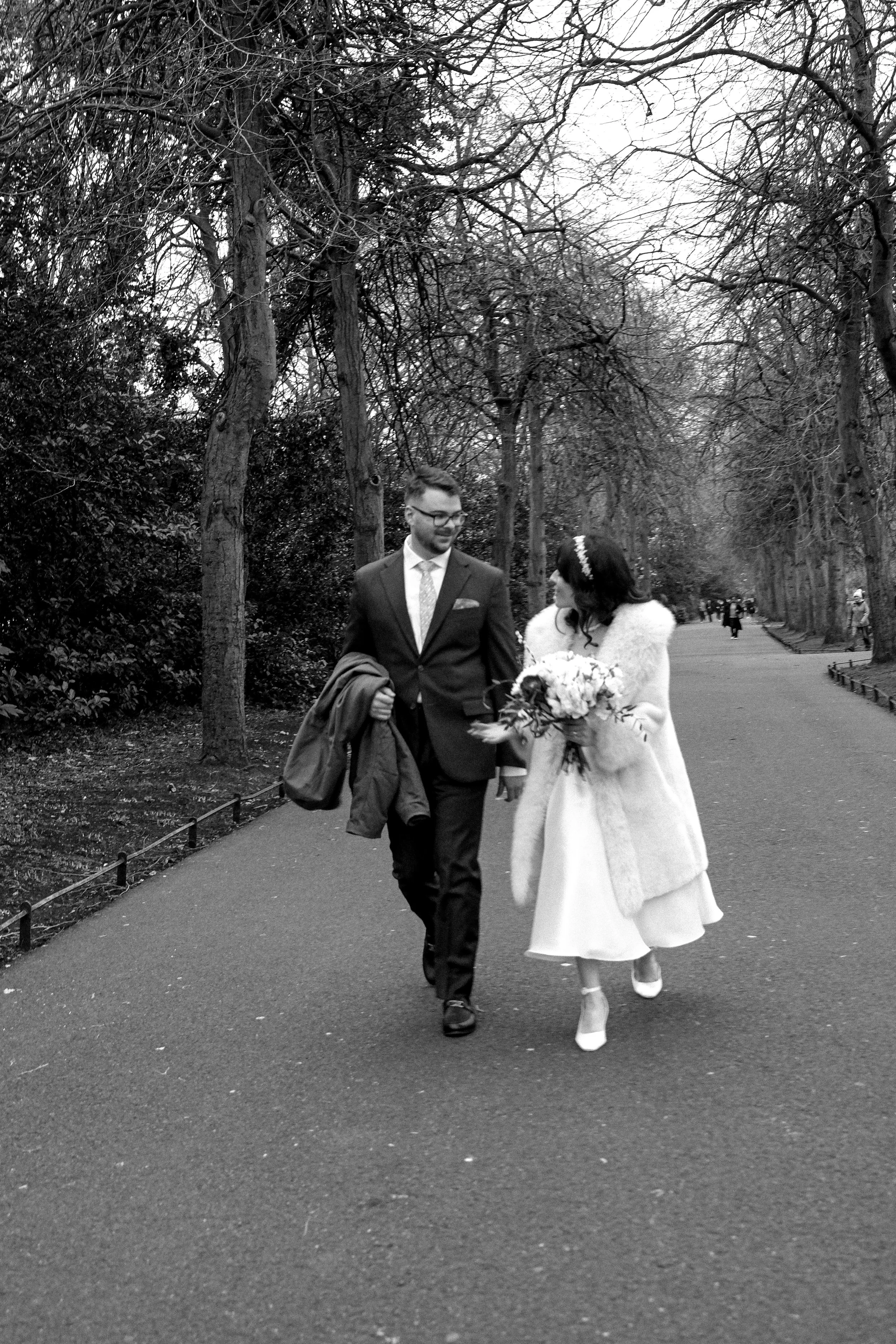 A black and white photo of a couple dressed in wedding attire walking together in a park, surrounded by leafless trees.