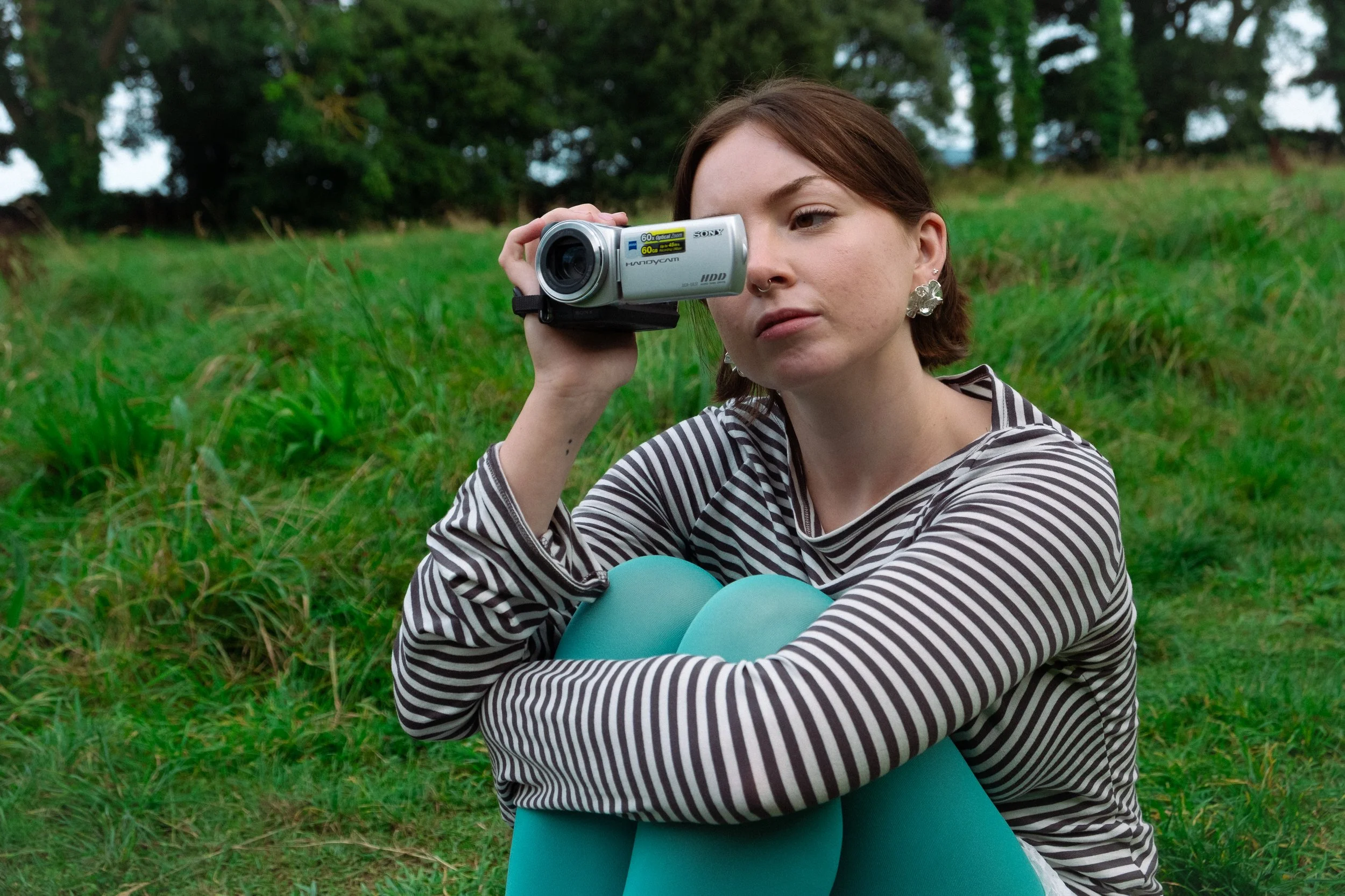 A young woman with short brown hair and earrings sitting on grass in a park, holding a video camera to her eye, wearing a striped long-sleeve shirt and turquoise leggings.