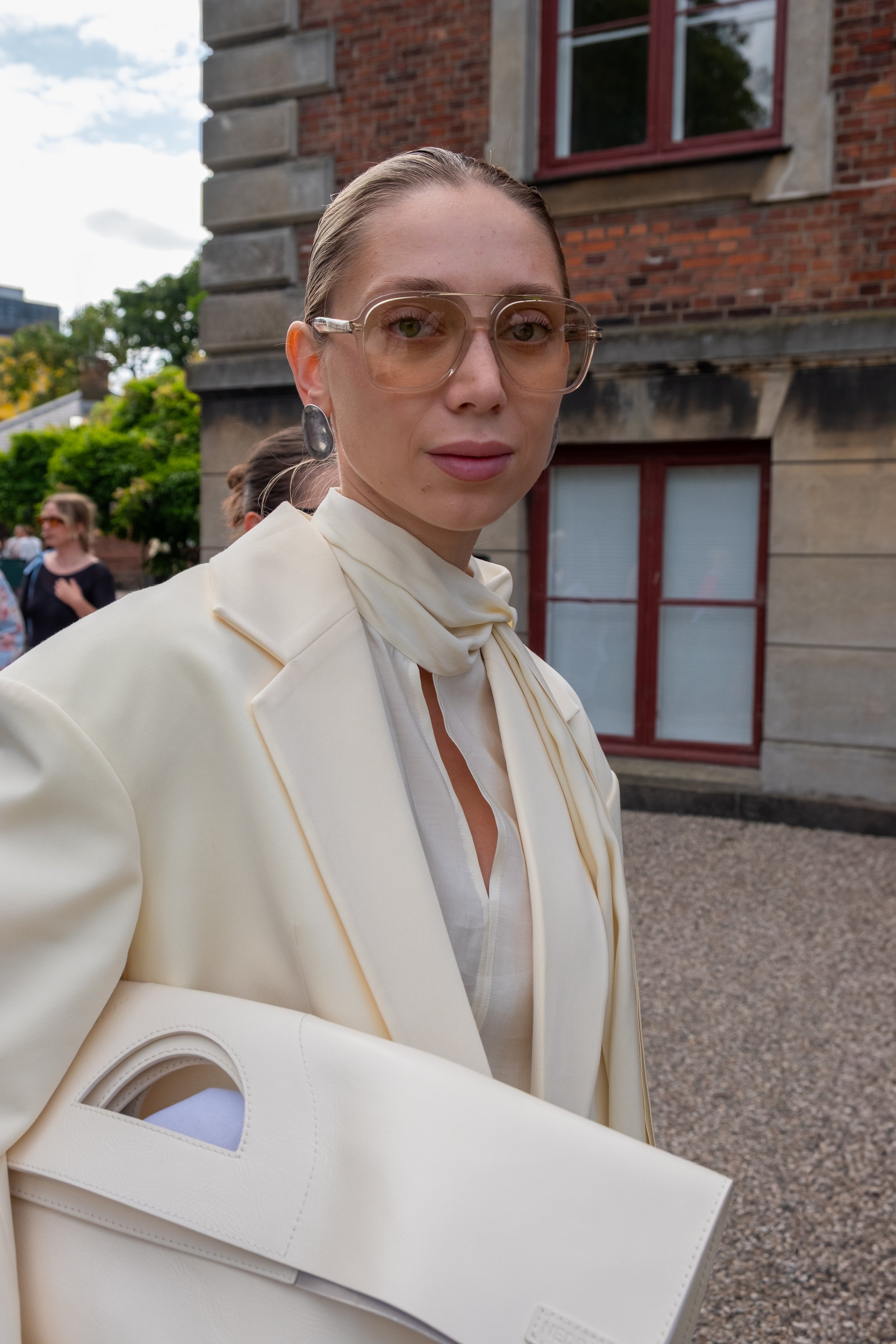 A woman with blonde hair tied back, wearing large tinted glasses, a cream-colored blazer, and a matching scarf, holding a large white handbag, standing outdoors in front of a brick building with other people in the background.