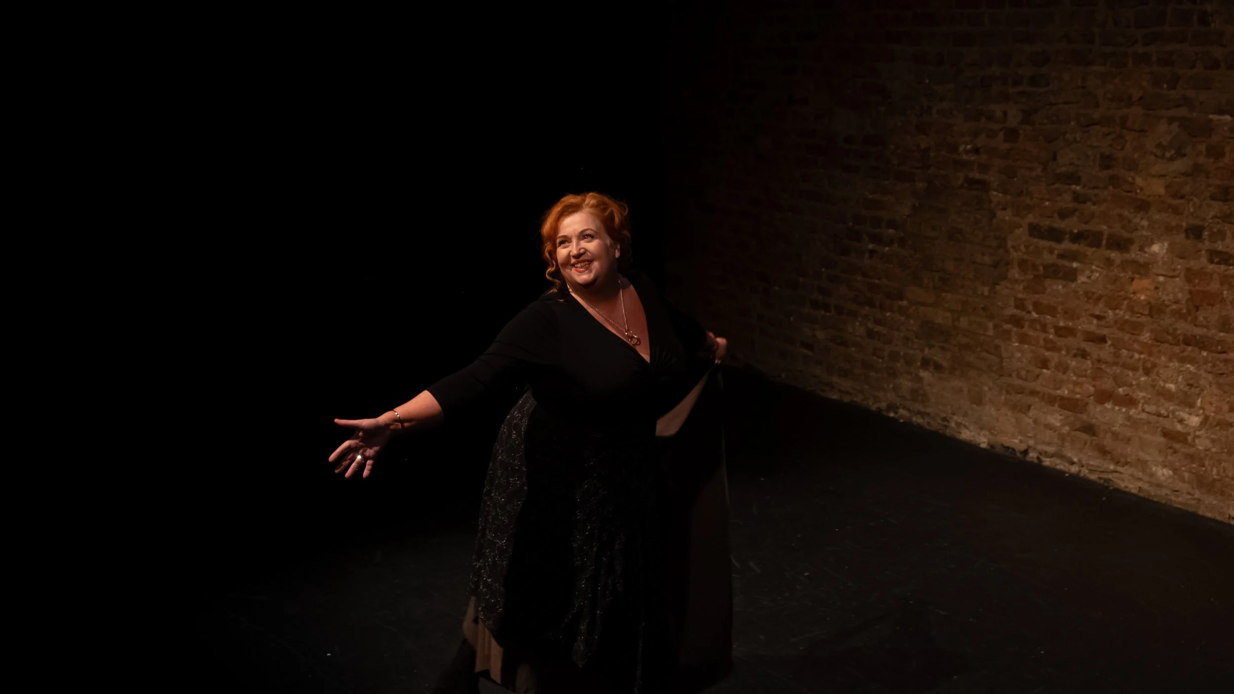 A smiling woman with red hair wearing a black dress, standing on a dark stage with a brick wall background.
