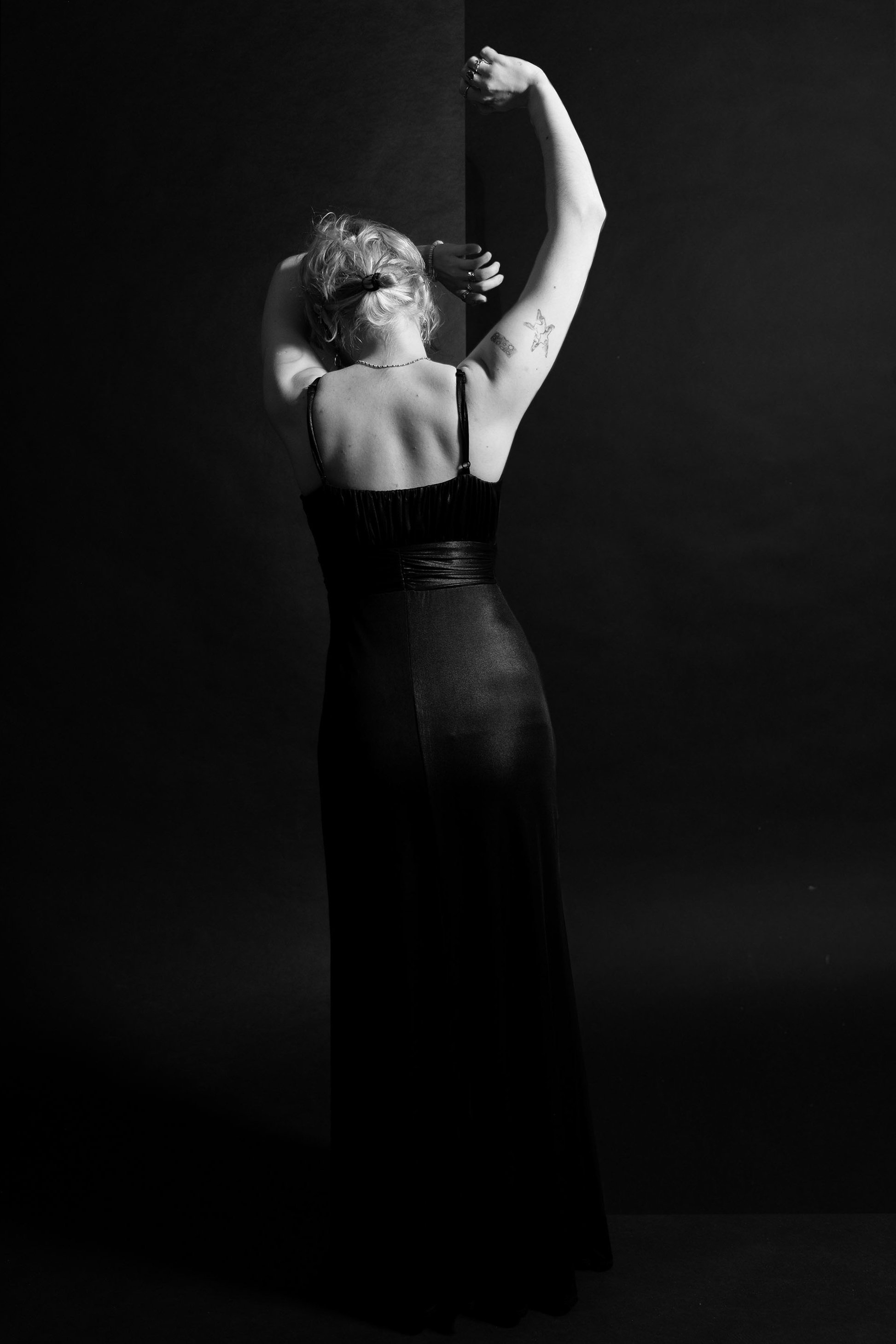 A woman in a black dress poses with her back to the camera, arms raised, against a dark background.
