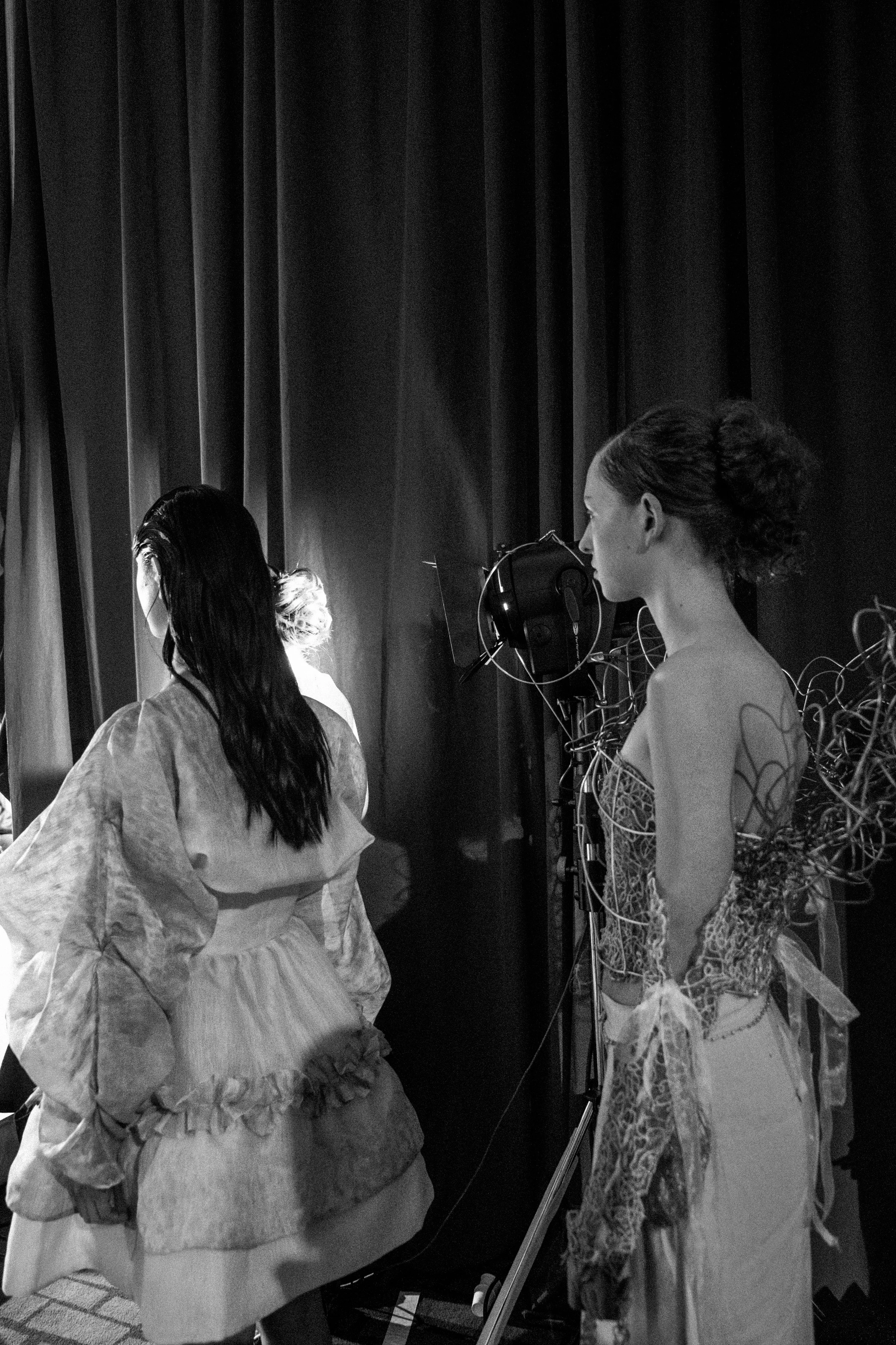 Two women in elaborate costumes standing behind stage curtains, preparing for a performance.