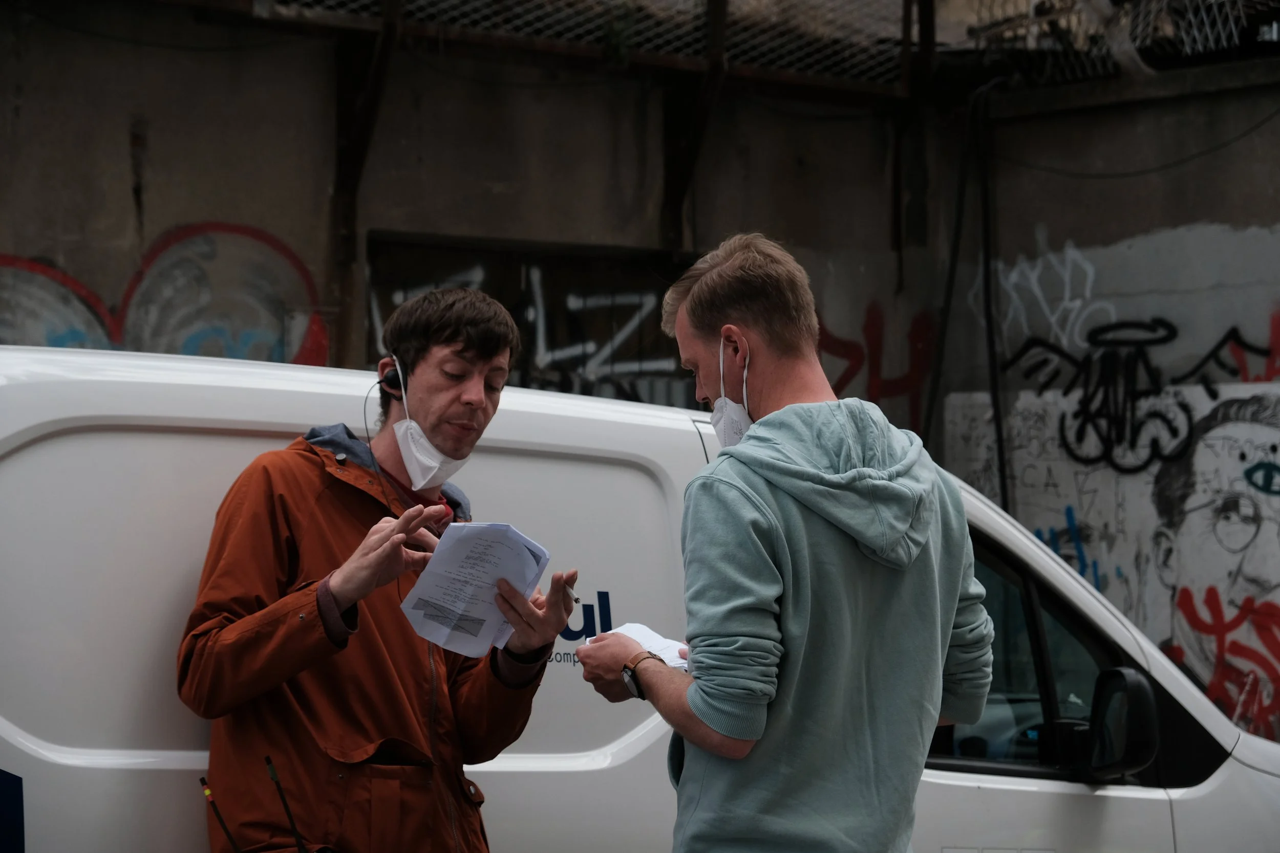 Two men wearing face masks and holding papers, standing beside a white van in an urban setting with graffiti-covered walls.