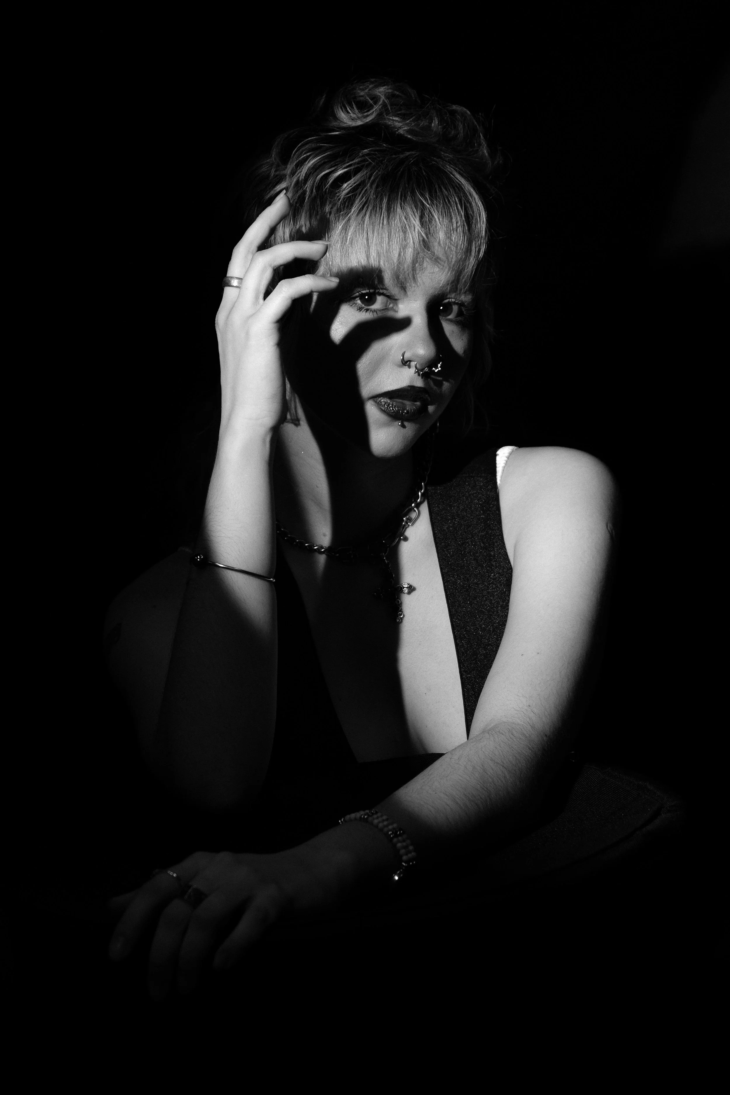 A woman with short, wavy hair and multiple facial piercings, wearing dark clothing and jewelry, poses against a dark background, illuminated by a stark light source.