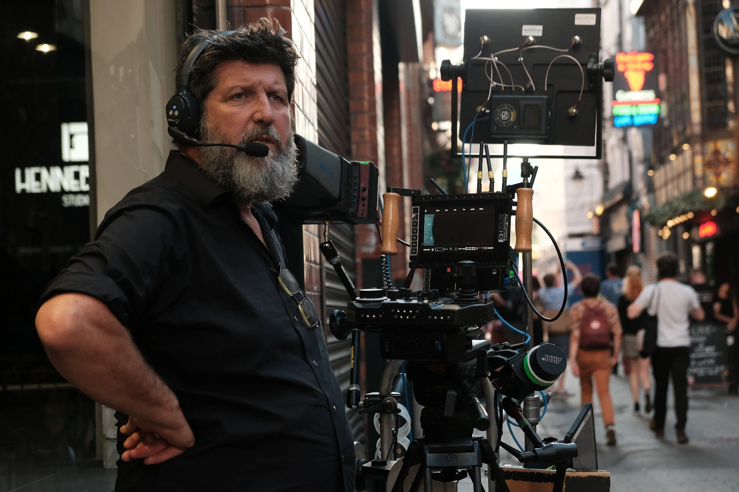 A man with a beard and curly hair wearing a black shirt and a headset operating a professional film camera on a city street with pedestrians in the background.