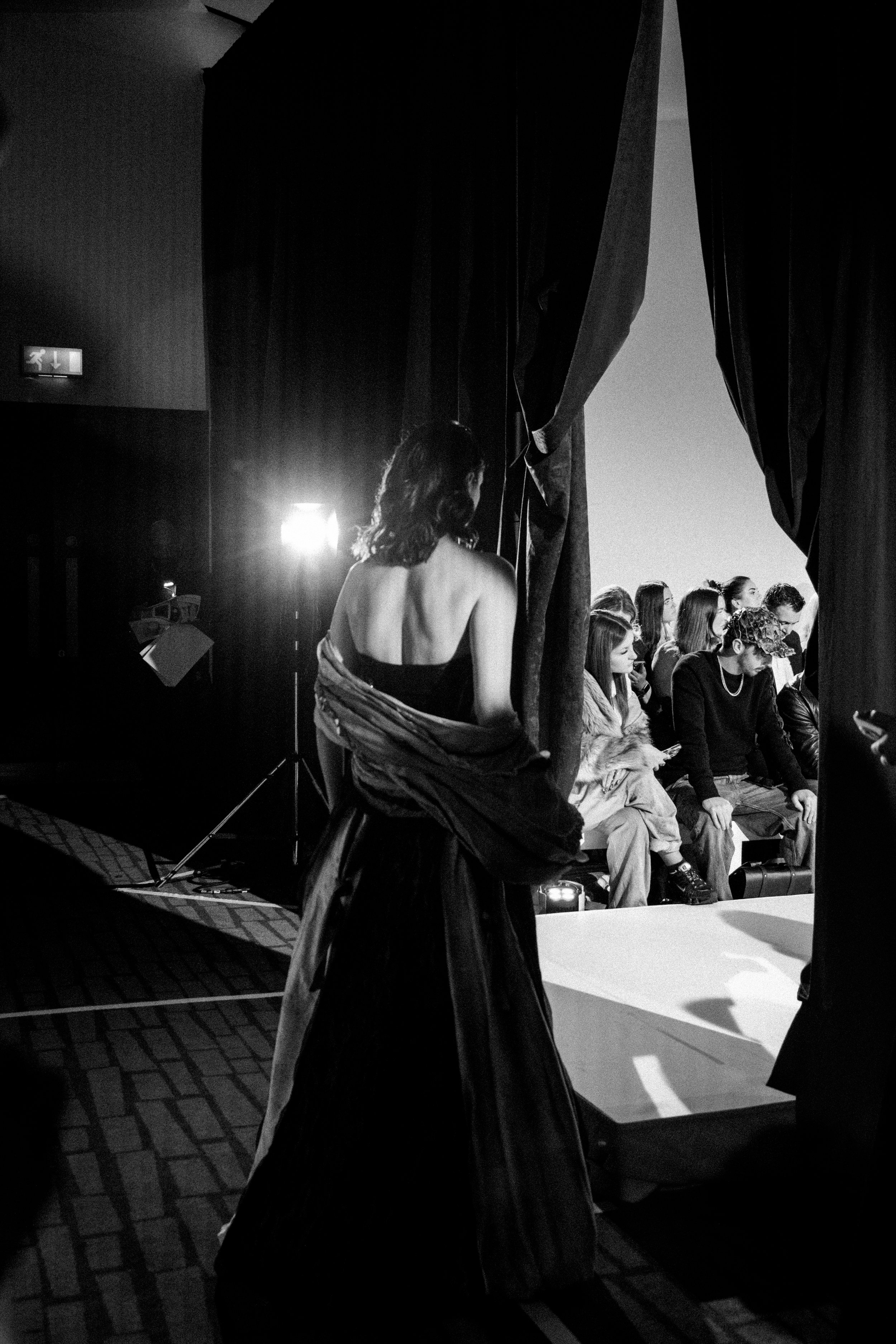 A woman in a strapless black dress is preparing to go on stage, seen from behind, at a fashion or runway show with seated audience members watching.