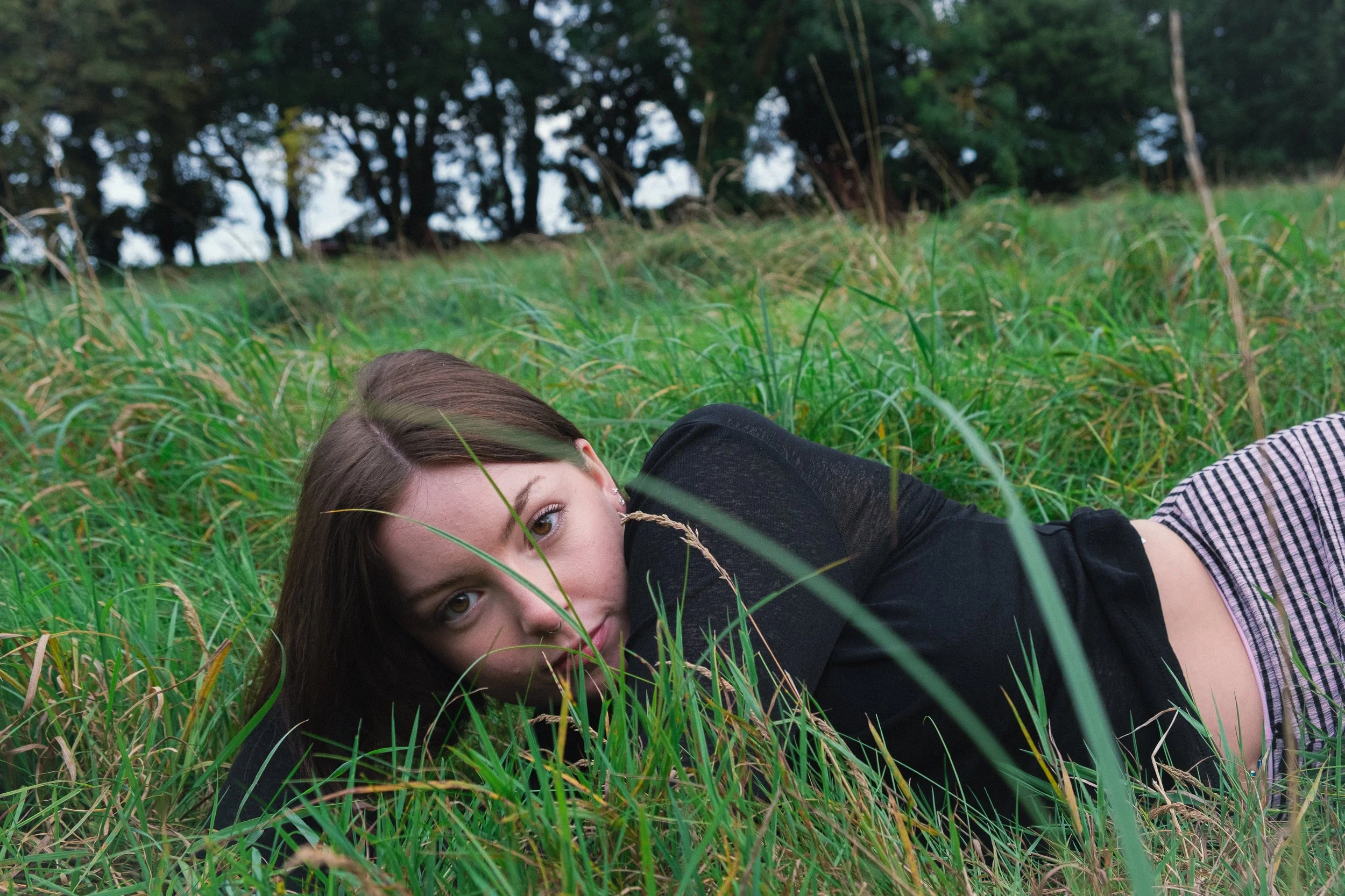 A young woman lying on her side in a field of tall grass with trees in the background, looking at the camera.