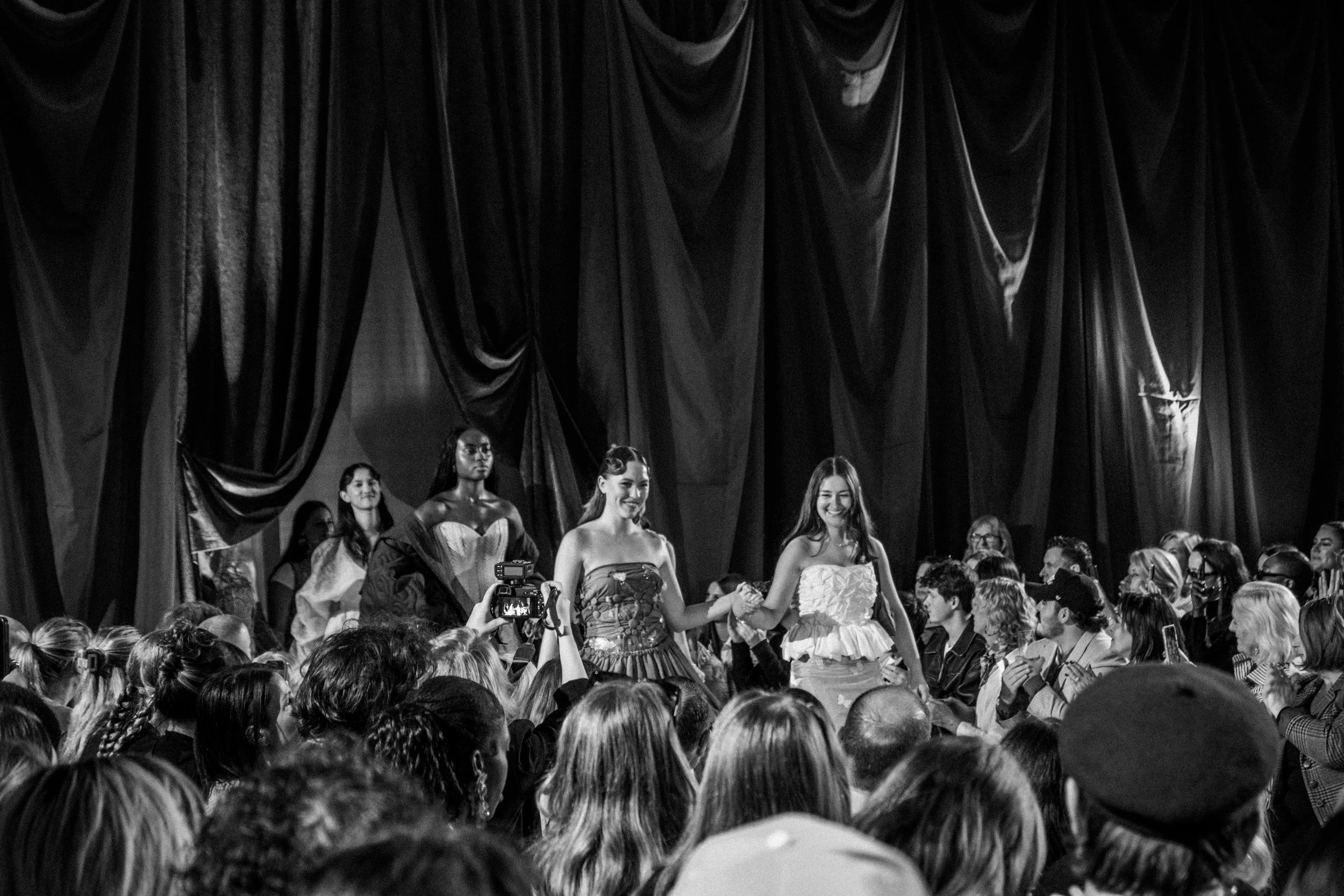Black and white photo of a fashion show with two models holding hands on the runway, surrounded by an audience.