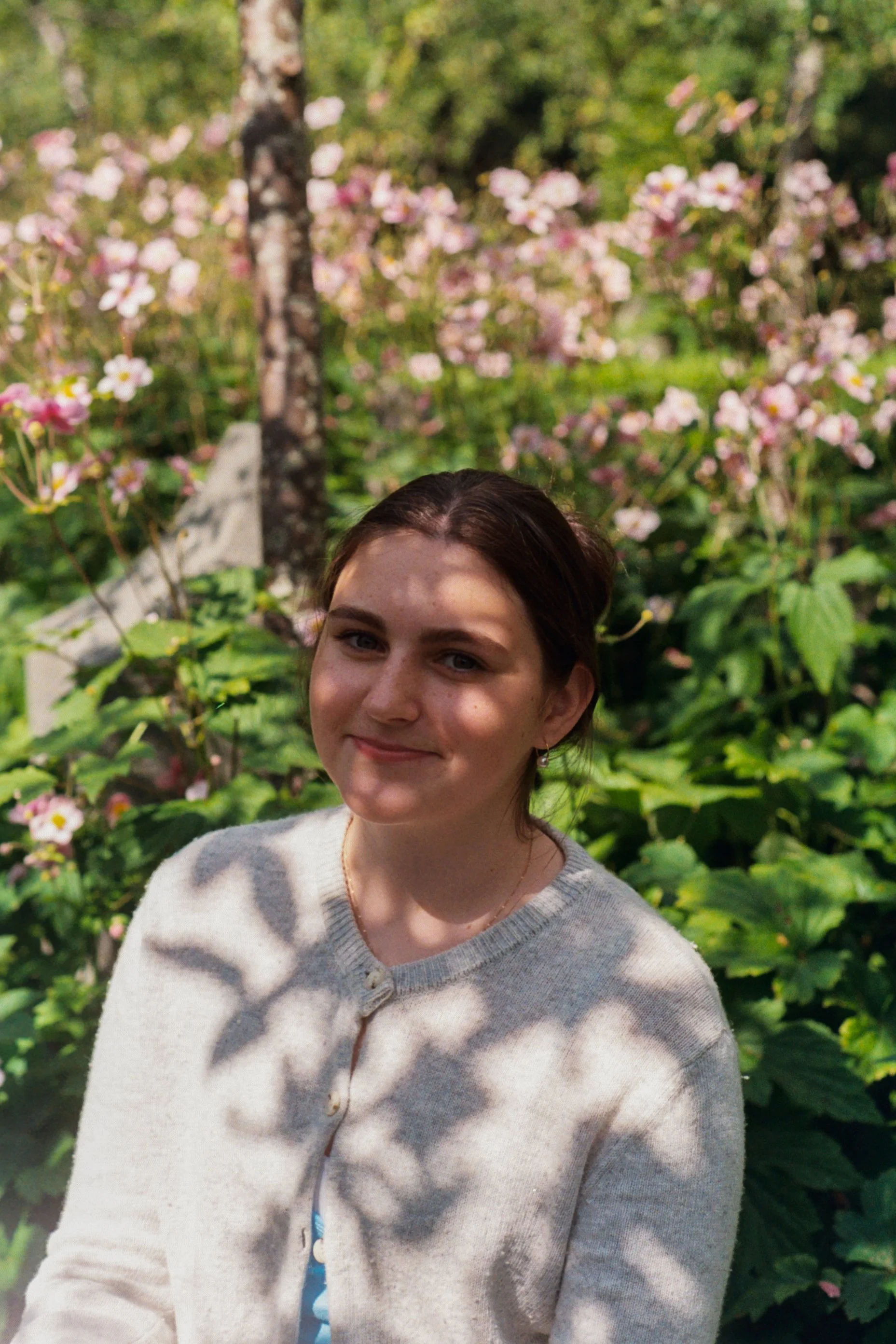 A young woman with dark brown hair and earrings smiling outdoors in front of pink and white flowers and green foliage.