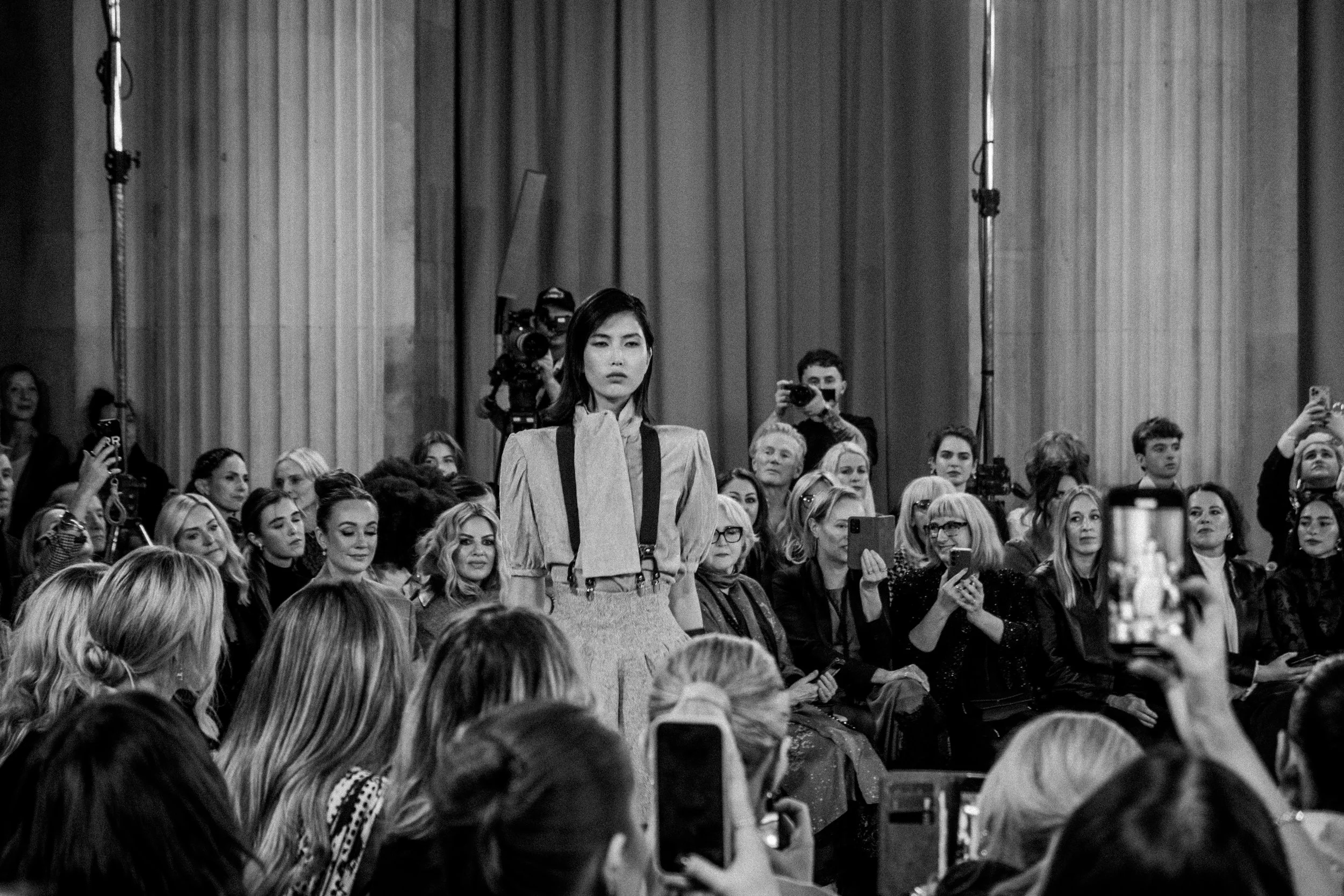 A woman on a runway during a fashion show, with an audience on both sides capturing the moment with their phones, taken in black and white.