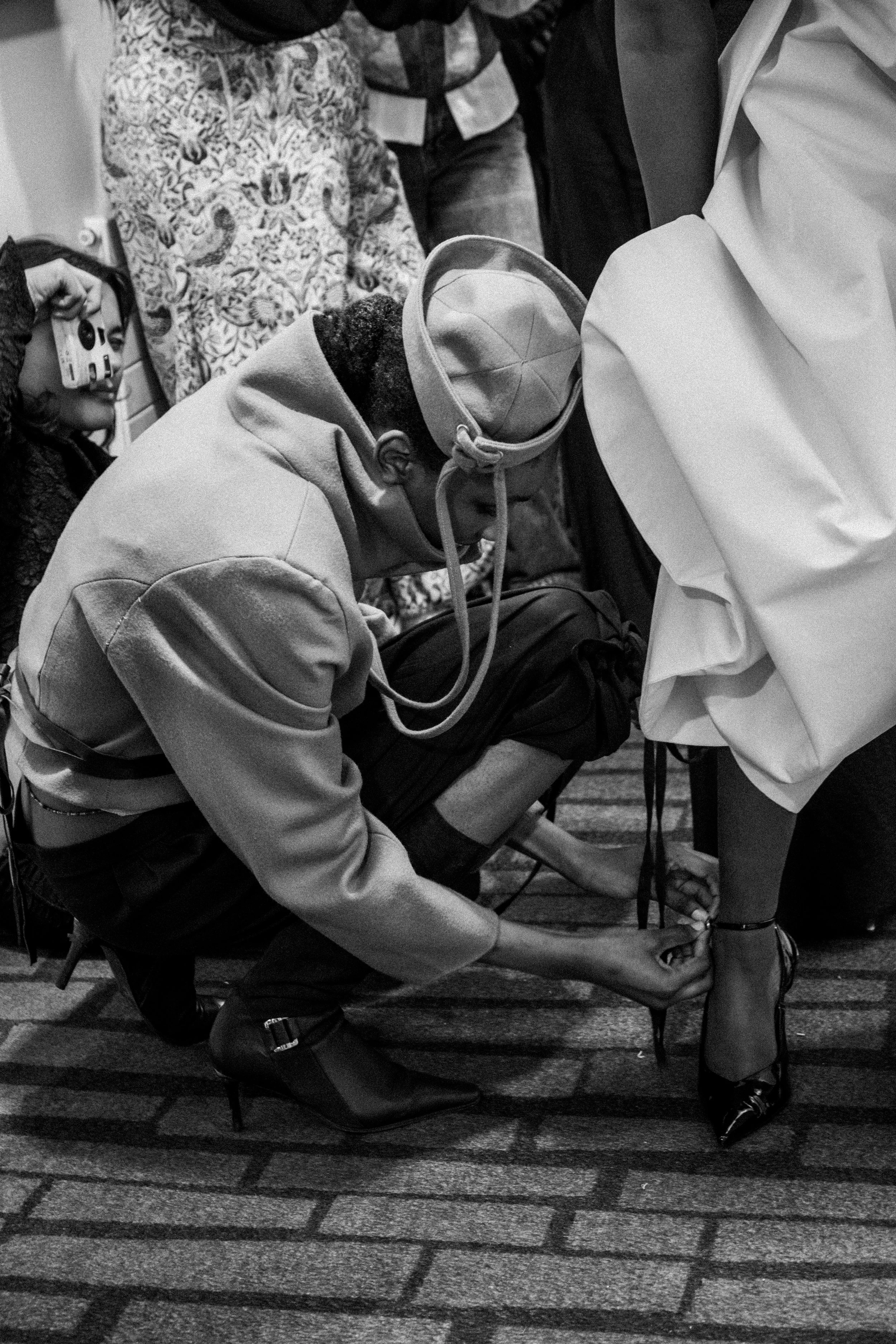 A man crouches down to tighten the strap on a woman's high heel shoe, while surrounded by other people in a crowded onyx. The man is wearing a hoodie, and the woman is wearing a white garment and patterned shoes.