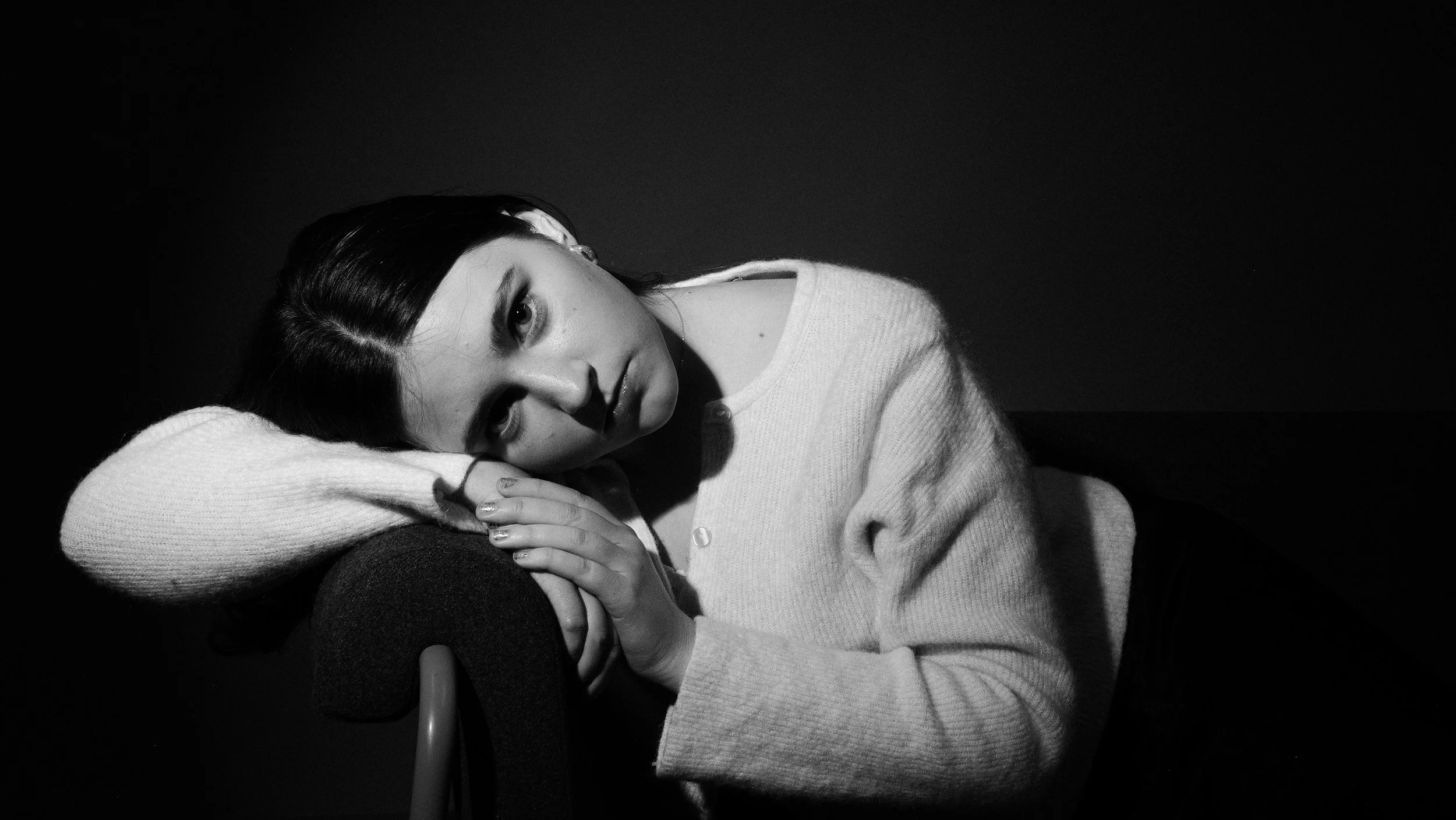 A black and white portrait of a woman resting her head on her arms, looking directly at the camera with a contemplative expression.