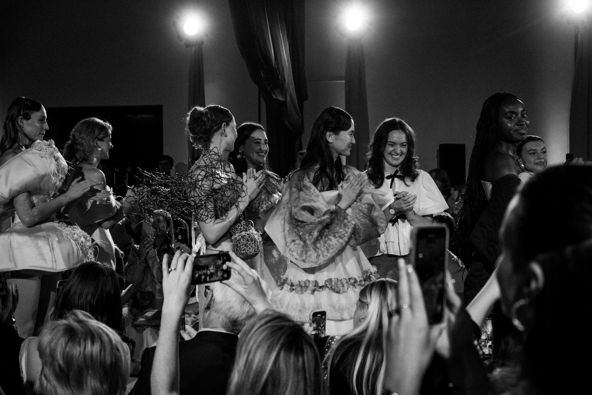 A group of women standing on stage at a celebration or event, smiling and clapping, with audience members taking photos in the foreground. The scene is lit with stage lights, and the women are dressed in elegant and traditional outfits.