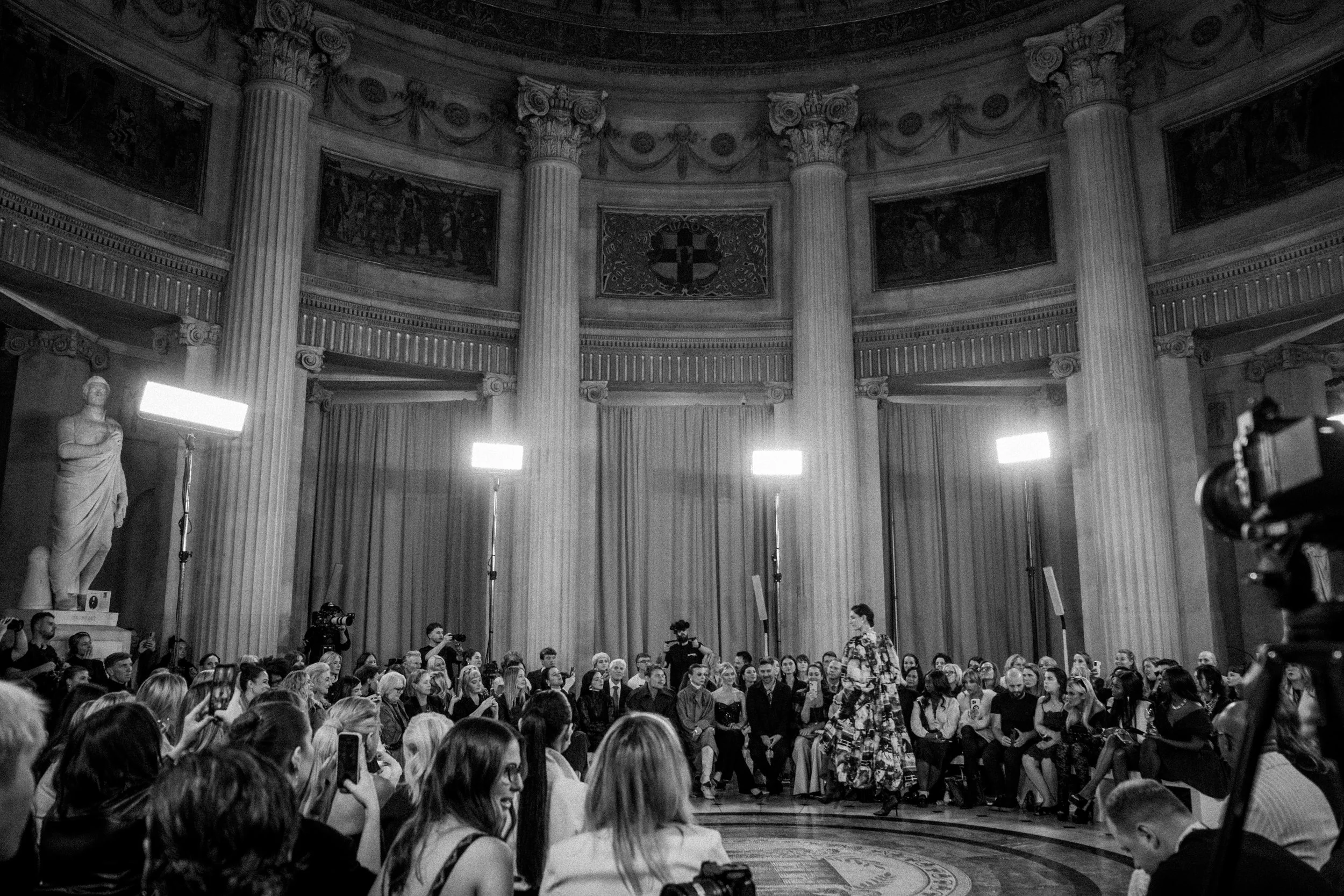 A fashion show in a grand, classical hall with large columns and statues, with models walking on the runway and seated audience watching.