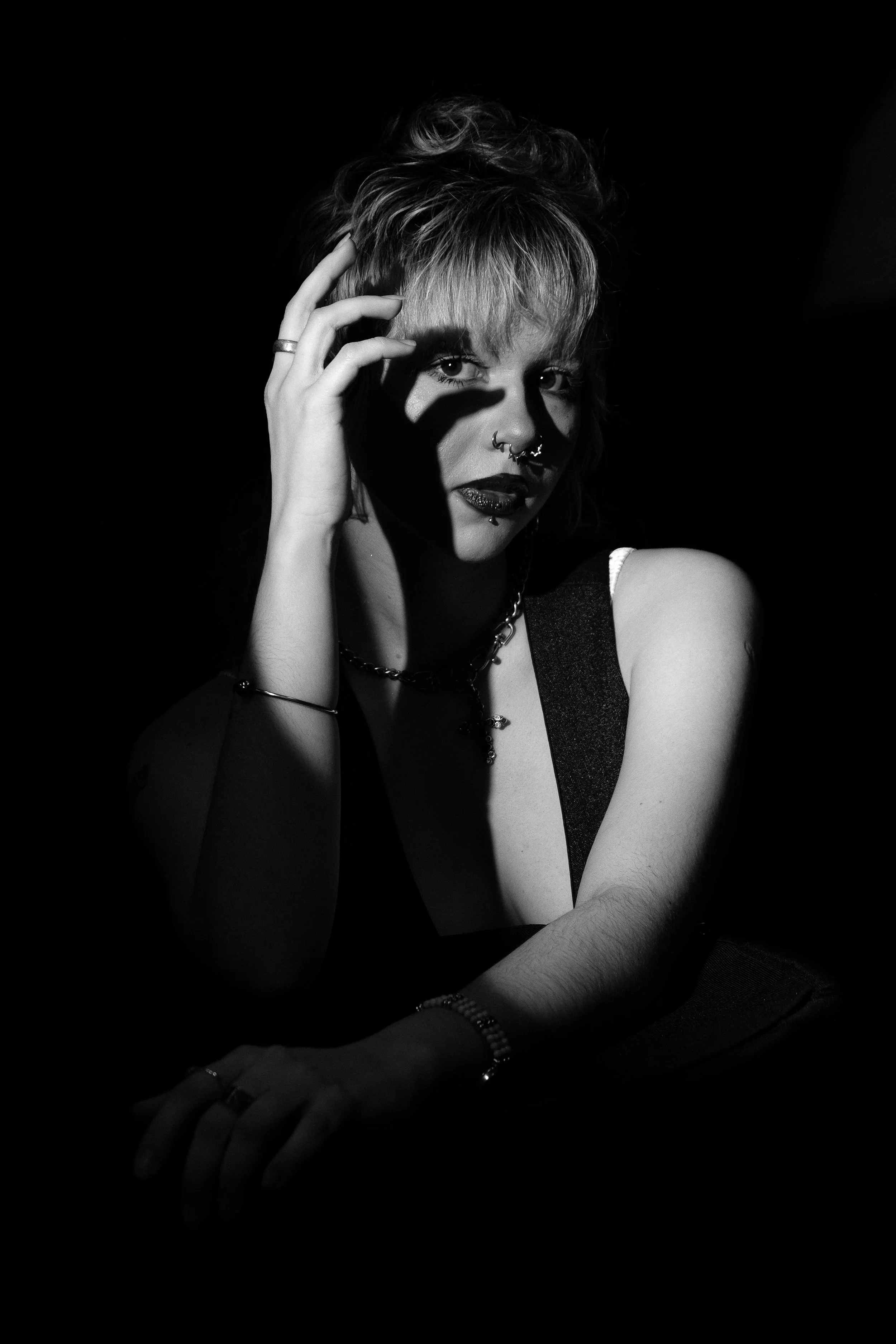 A black and white photo of a woman with short hair, multiple facial piercings, and dark lipstick, posing with her hand near her face against a dark background.