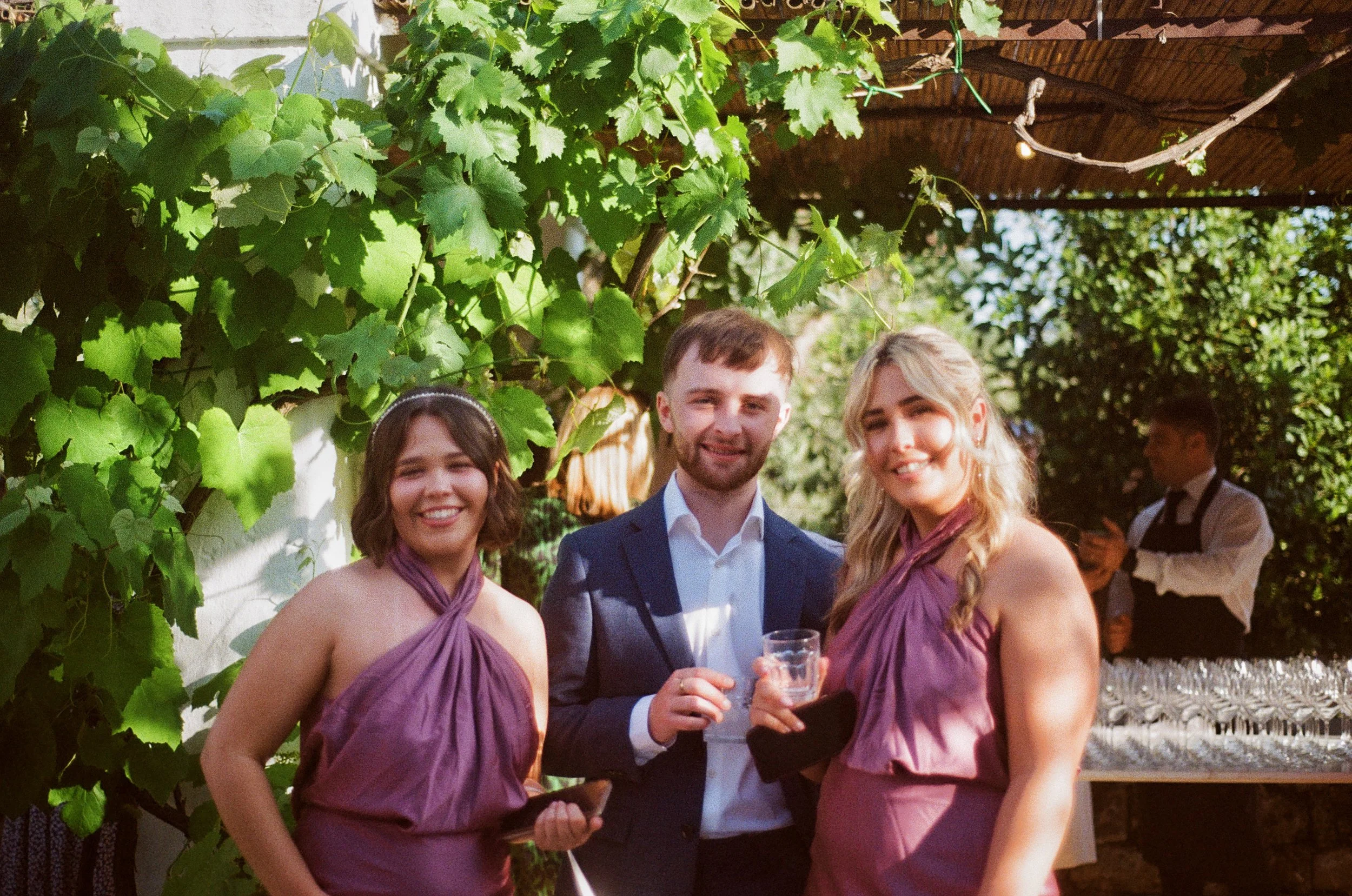 Three young adults smiling and holding drinks at an outdoor event, with greenery and a food or drink station in the background.