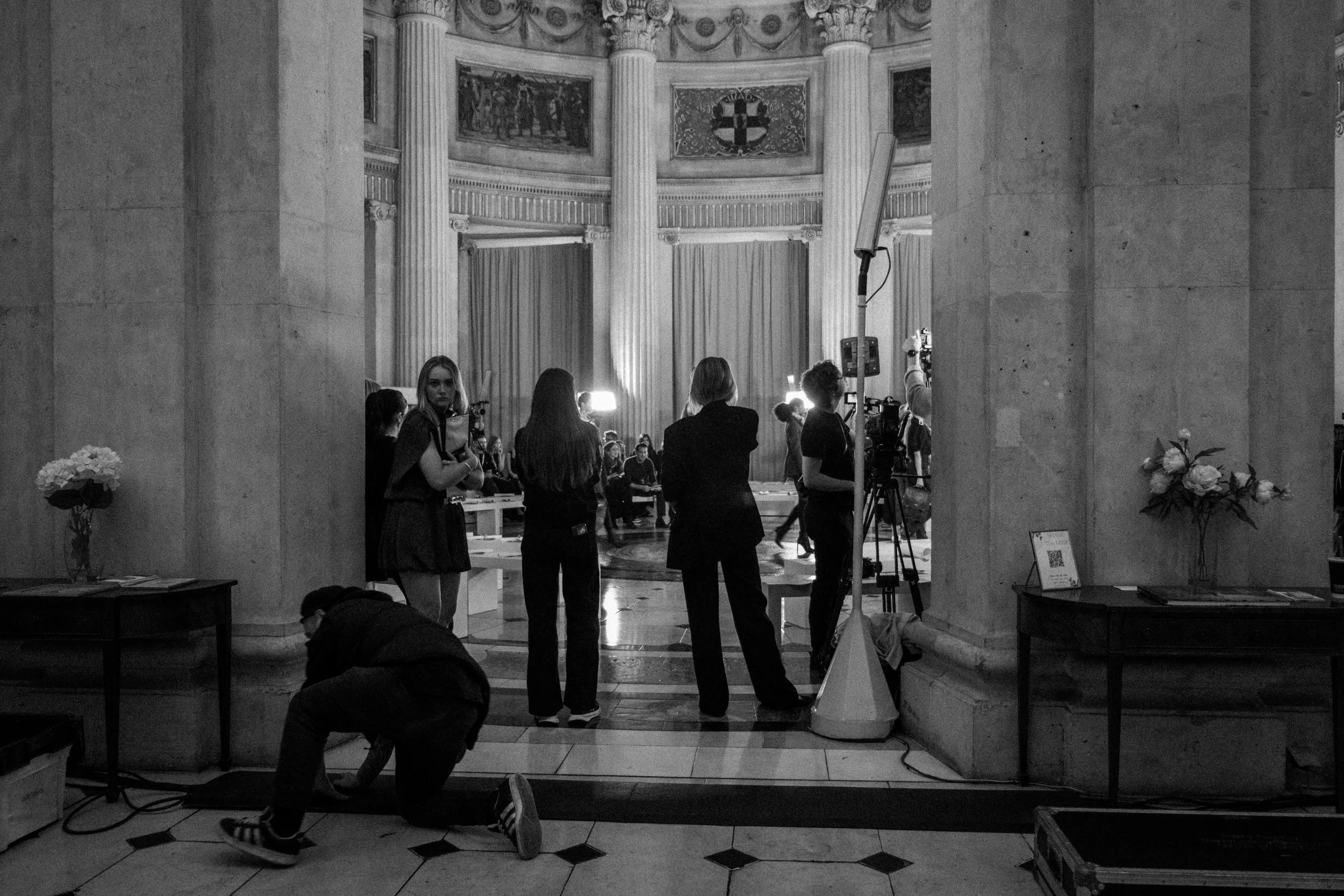 A black and white photo shows a group of people inside a large, ornate hall with tall columns, paintings, and decorative details. Some individuals are standing, possibly preparing for a interview or filming, with filming equipment and lighting set up