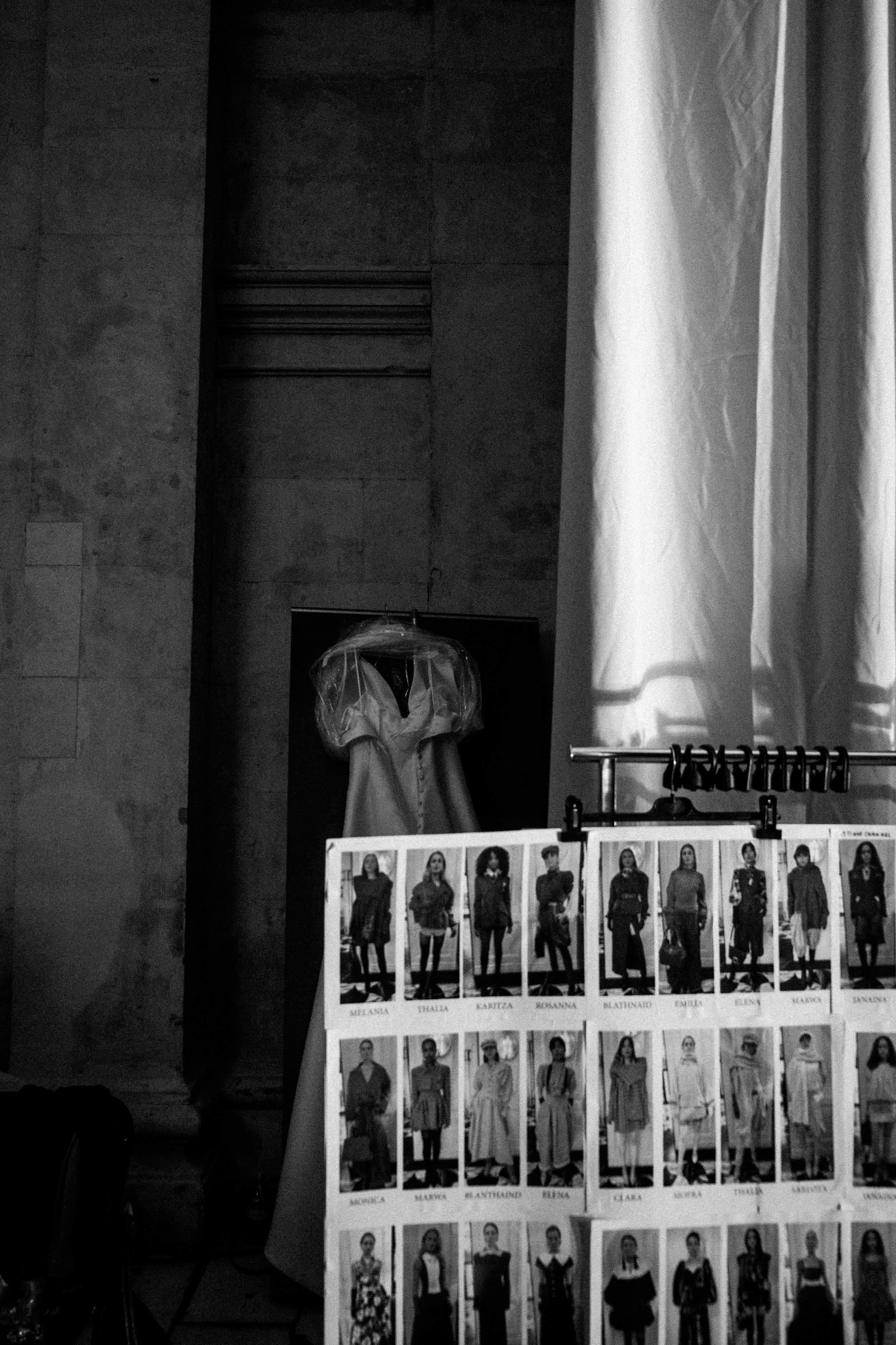 Black and white photo showing a clothing display at a fashion event, with a mannequin wearing a dress in the background and a board of model photos in the foreground.