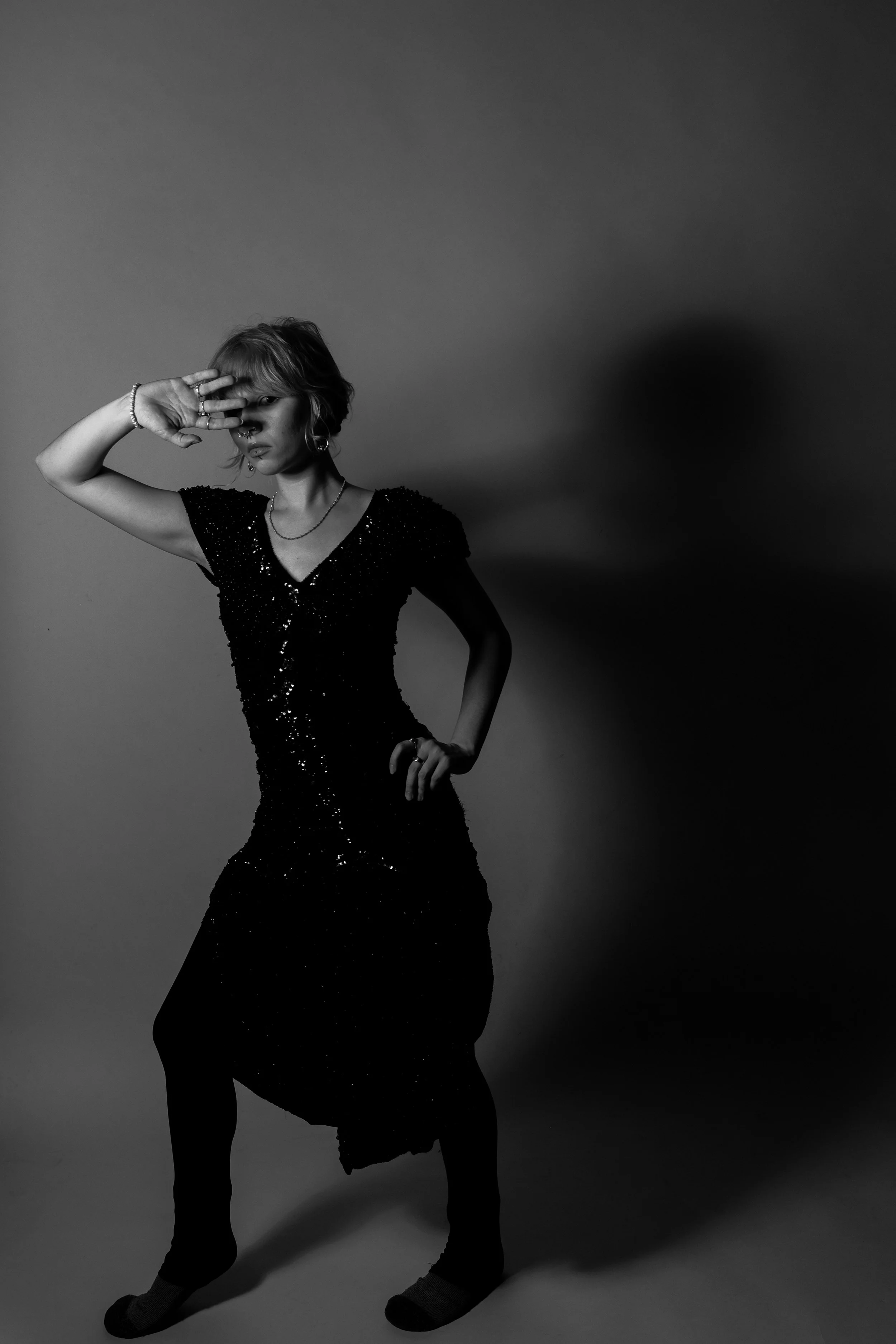 A woman in a black, sequined dress posing with one hand on her hip and the other covering part of her face, standing against a plain background.