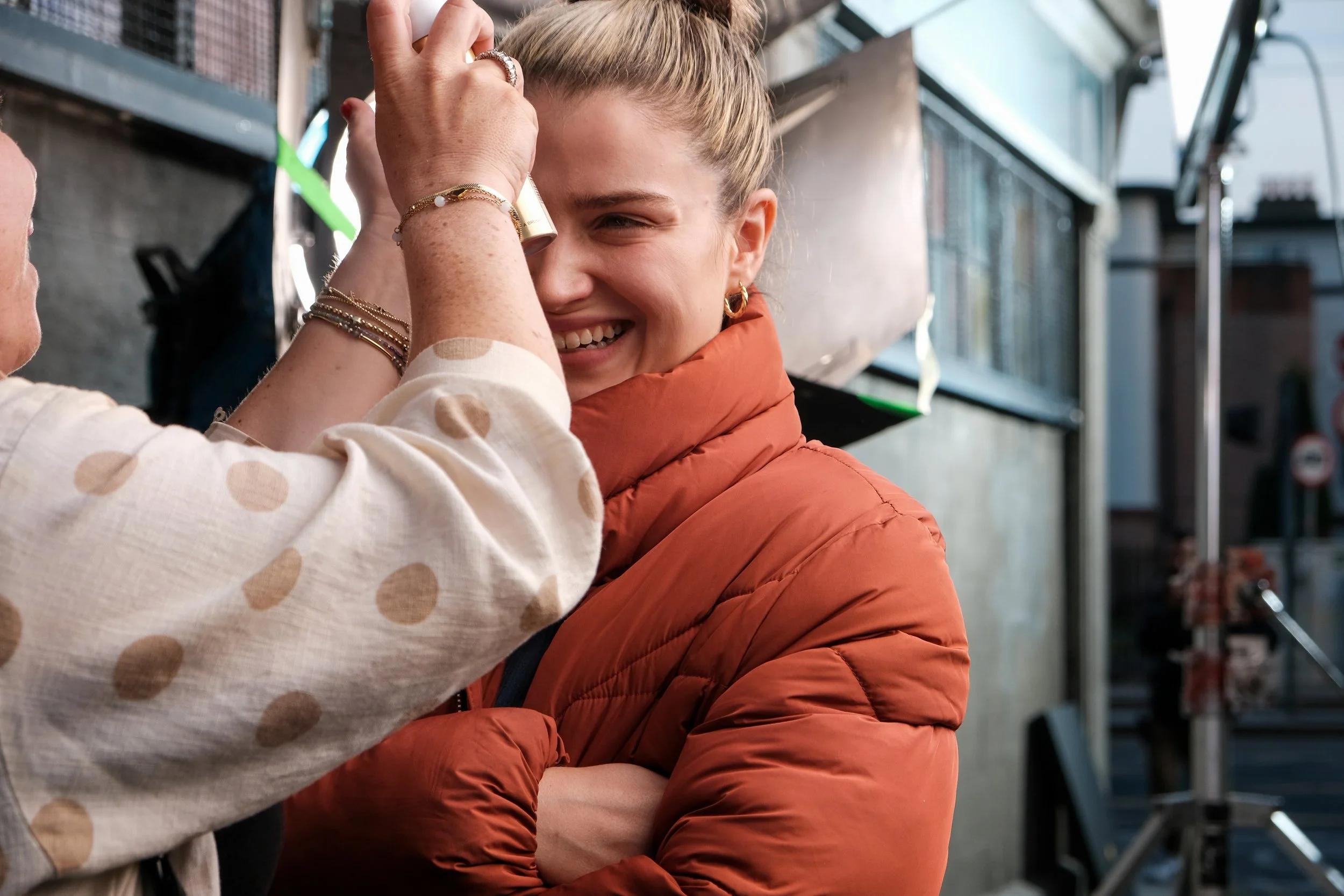 A woman in a white shirt with tan polka dots is applying makeup to a smiling woman wearing an orange jacket.