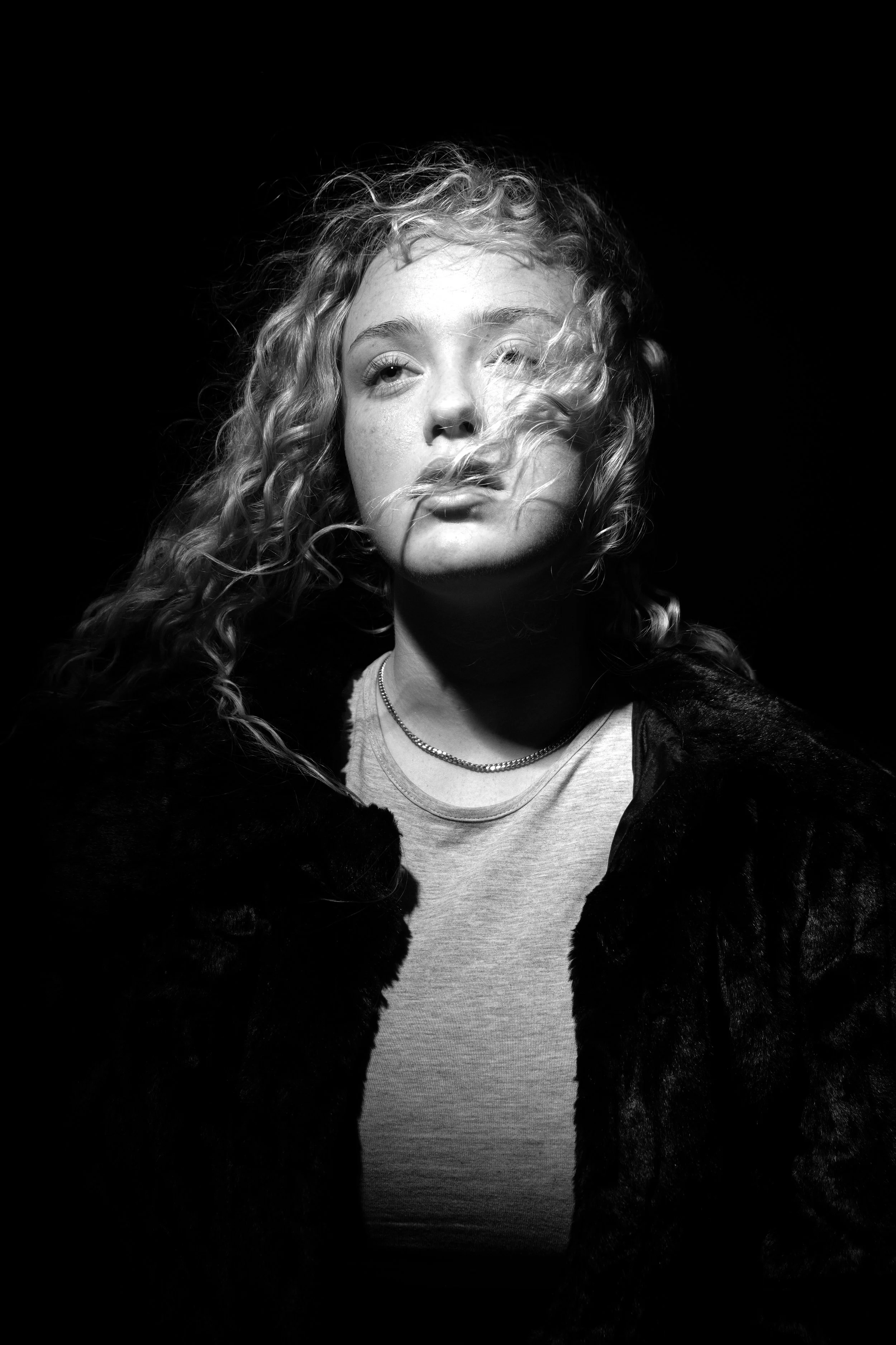 Black and white portrait of a young woman with curly hair partially covering her face, wearing a fur-like coat and a light-colored shirt, looking contemplative against a dark background.
