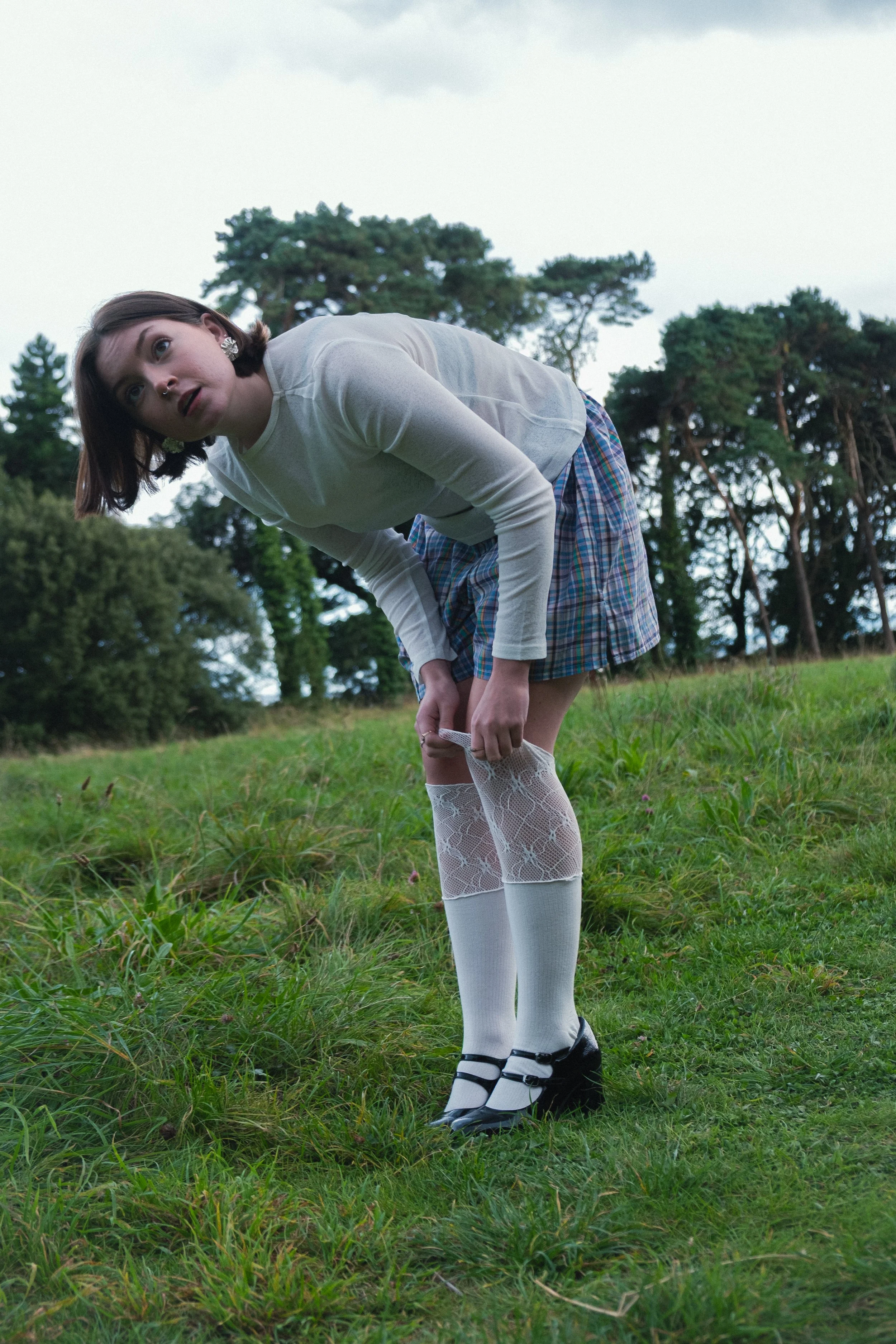 A woman in a white long-sleeve shirt, plaid skirt, and white thigh-high stockings with lace pattern, standing outdoors in a grassy area with trees in the background, bending over and looking to the side.