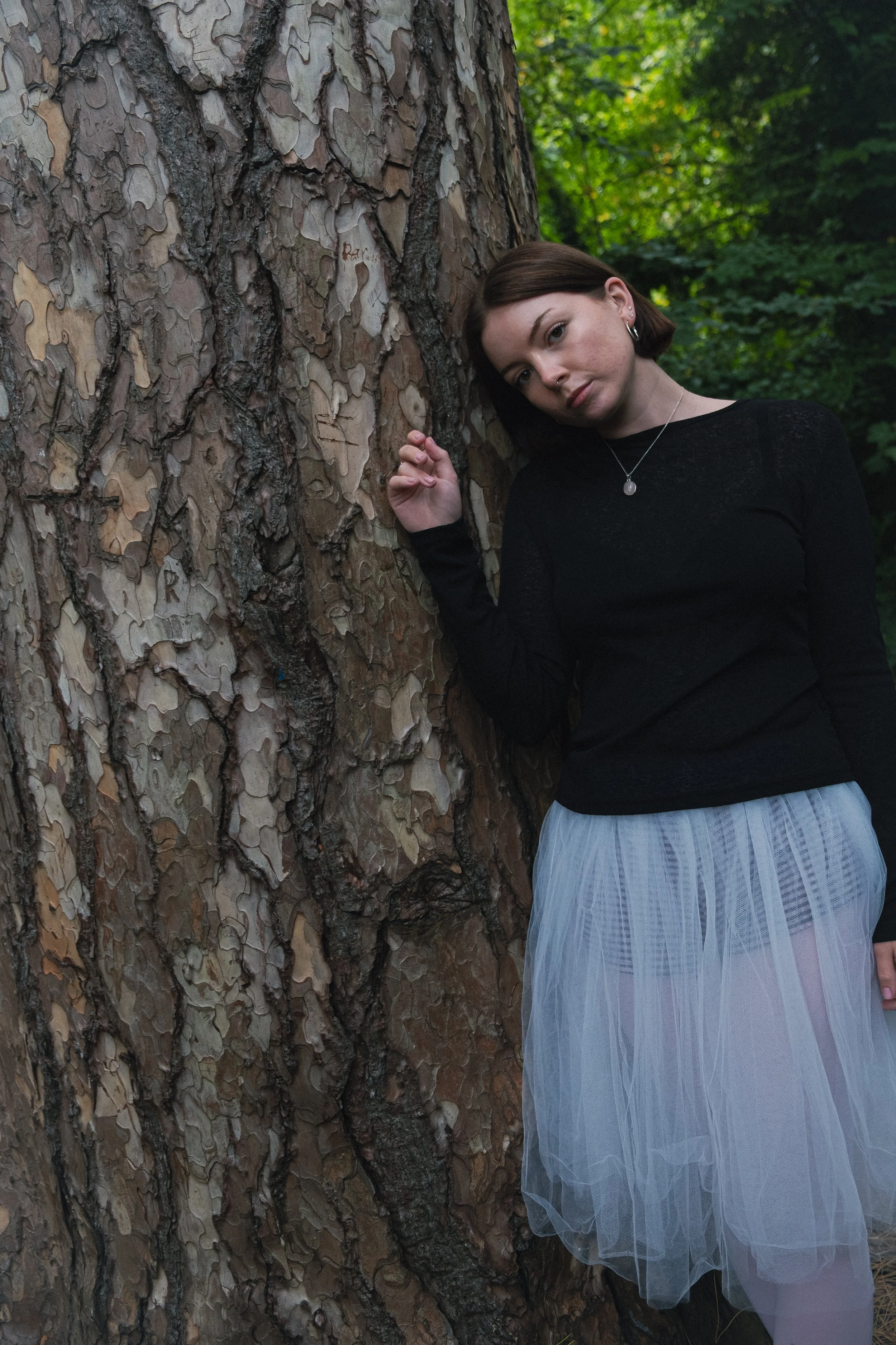 A young woman with brown hair and earrings leans against a large tree with textured bark, looking at the camera, in a wooded area.